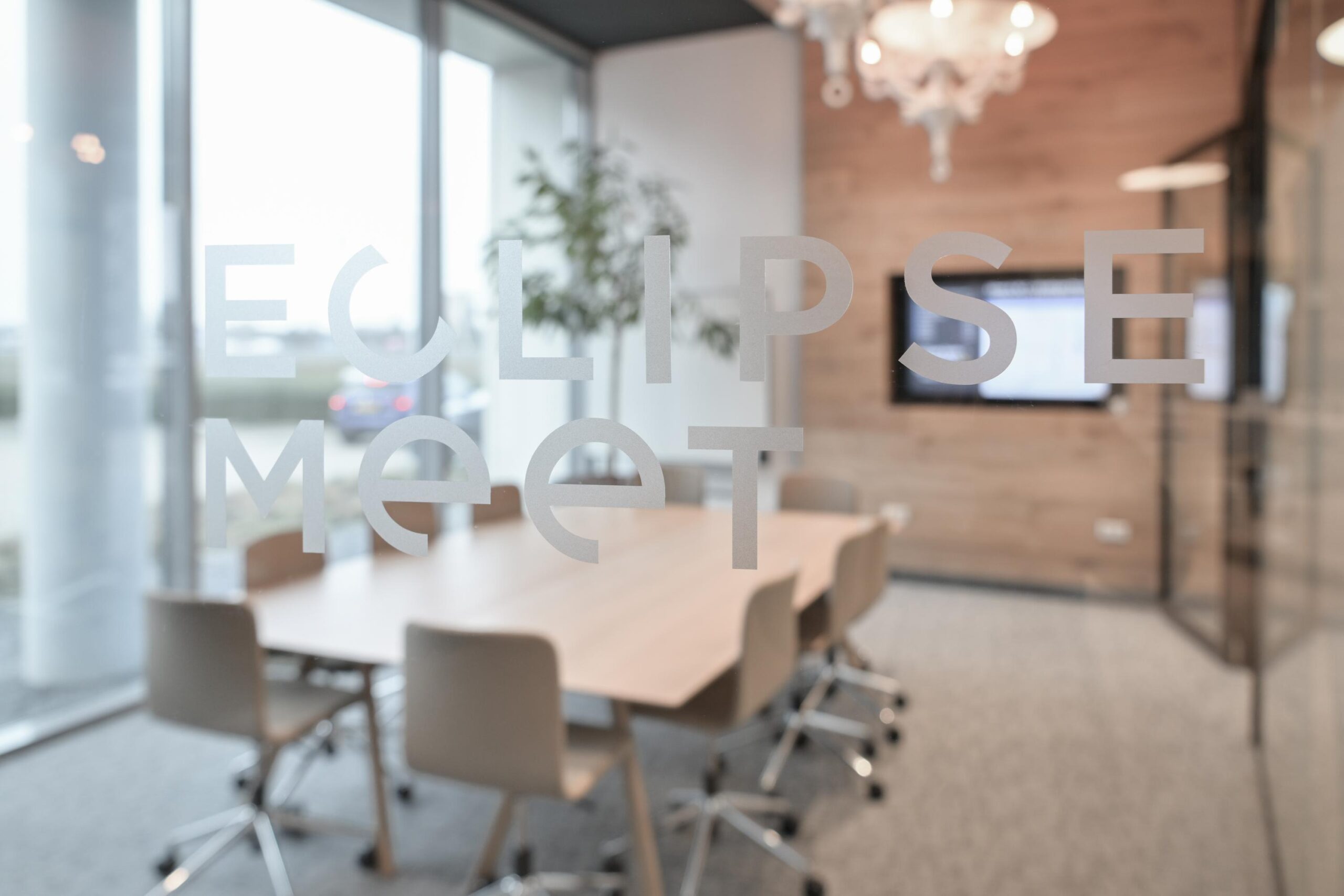 Vergaderruimte met een glazen wand waarop "Eclipse Meet" staat, en een houten tafel met stoelen.
