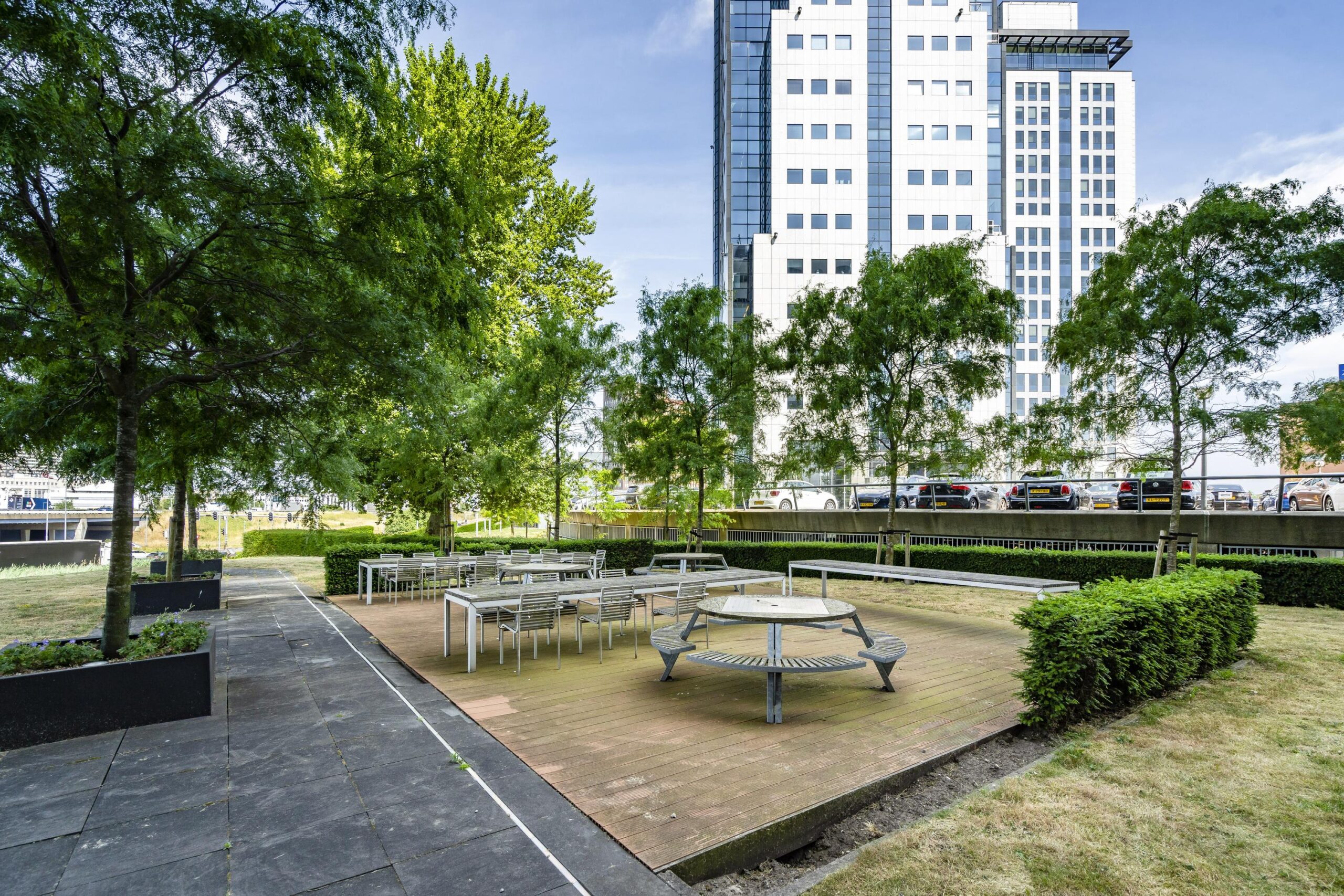 Buitenterras met tafels en banken bij Rivium Quadrant, omgeven door bomen en gras, met een hoog gebouw op de achtergrond.