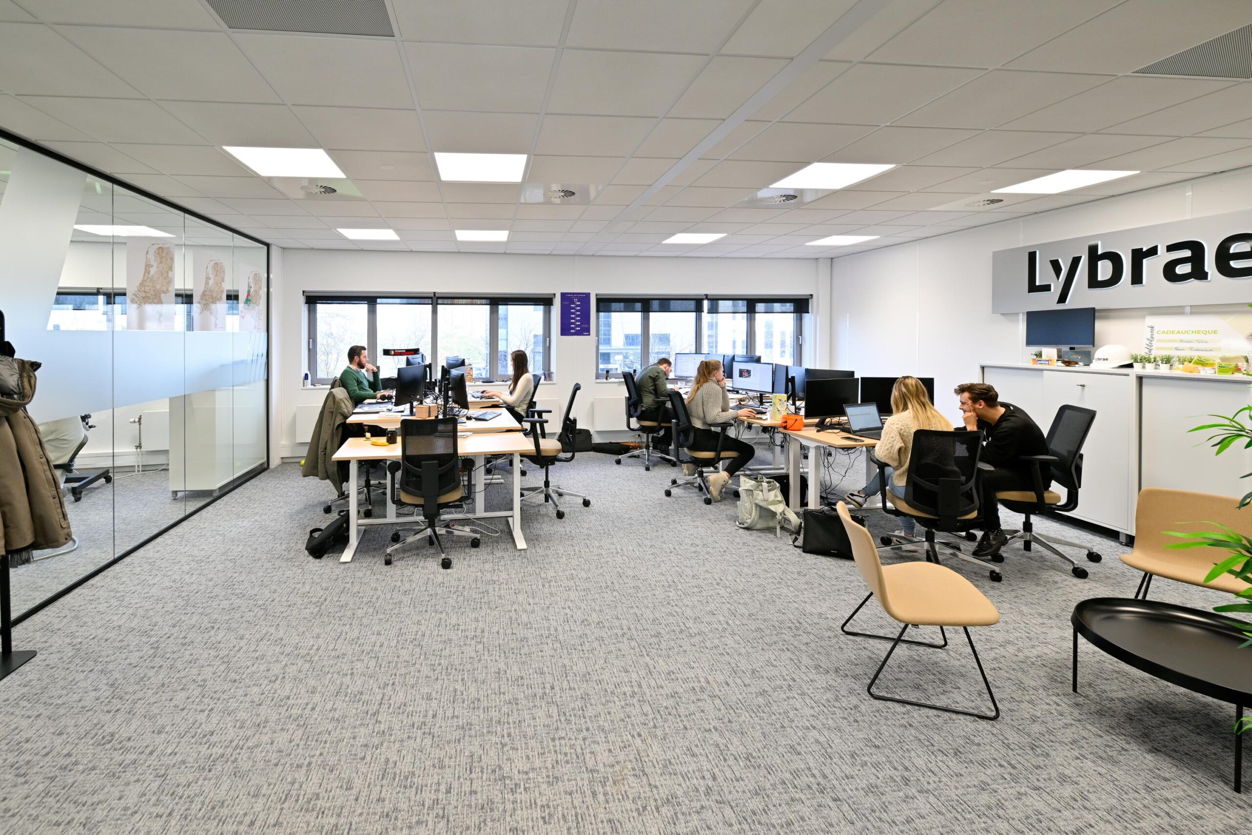 Een modern kantoor met meerdere mensen die aan bureaus werken in een open ruimte met de naam "Lybrae" aan de muur.