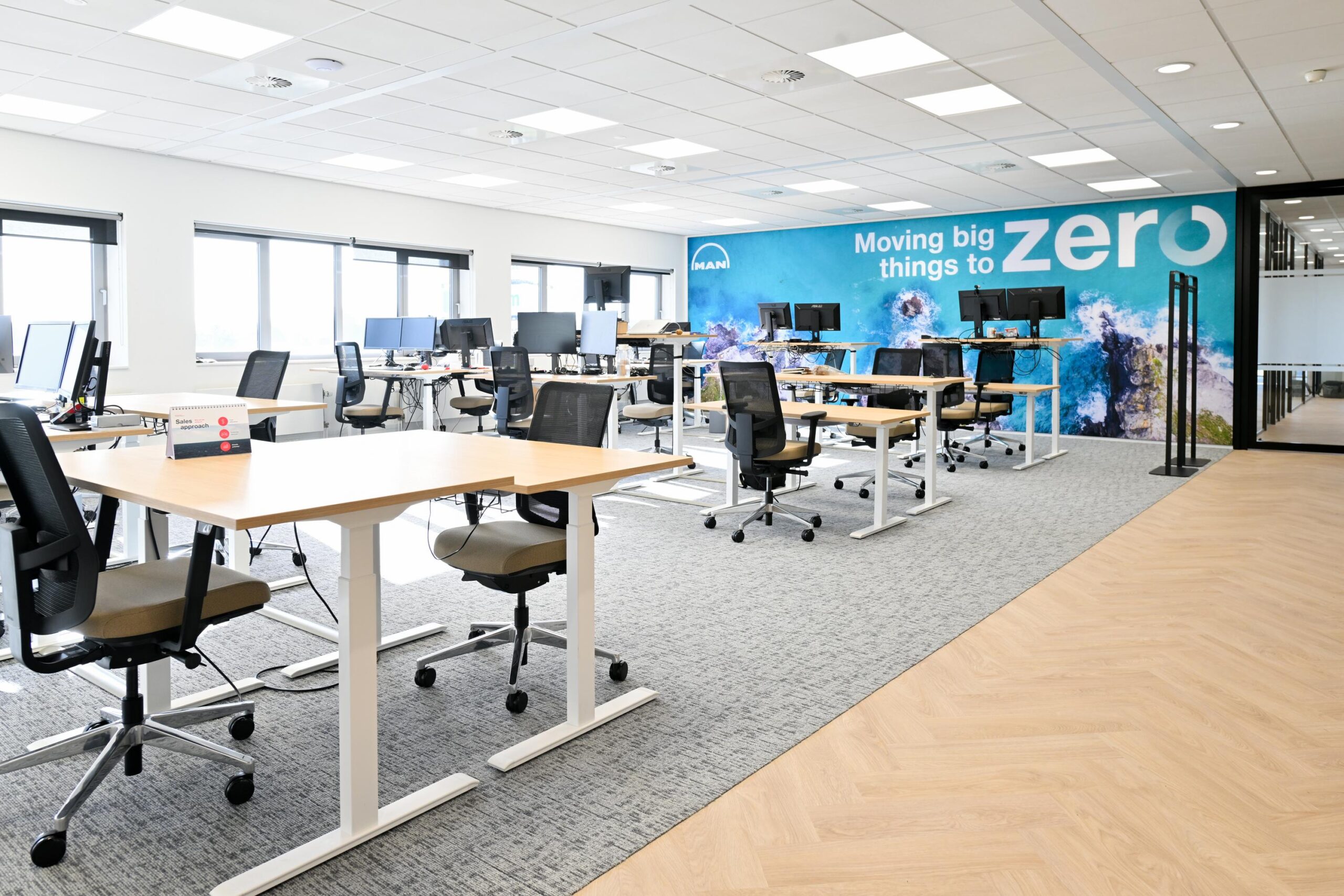 Een moderne, lichte kantoorruimte met bureaus, bureaustoelen en een wand met de tekst "Moving big things to zero".