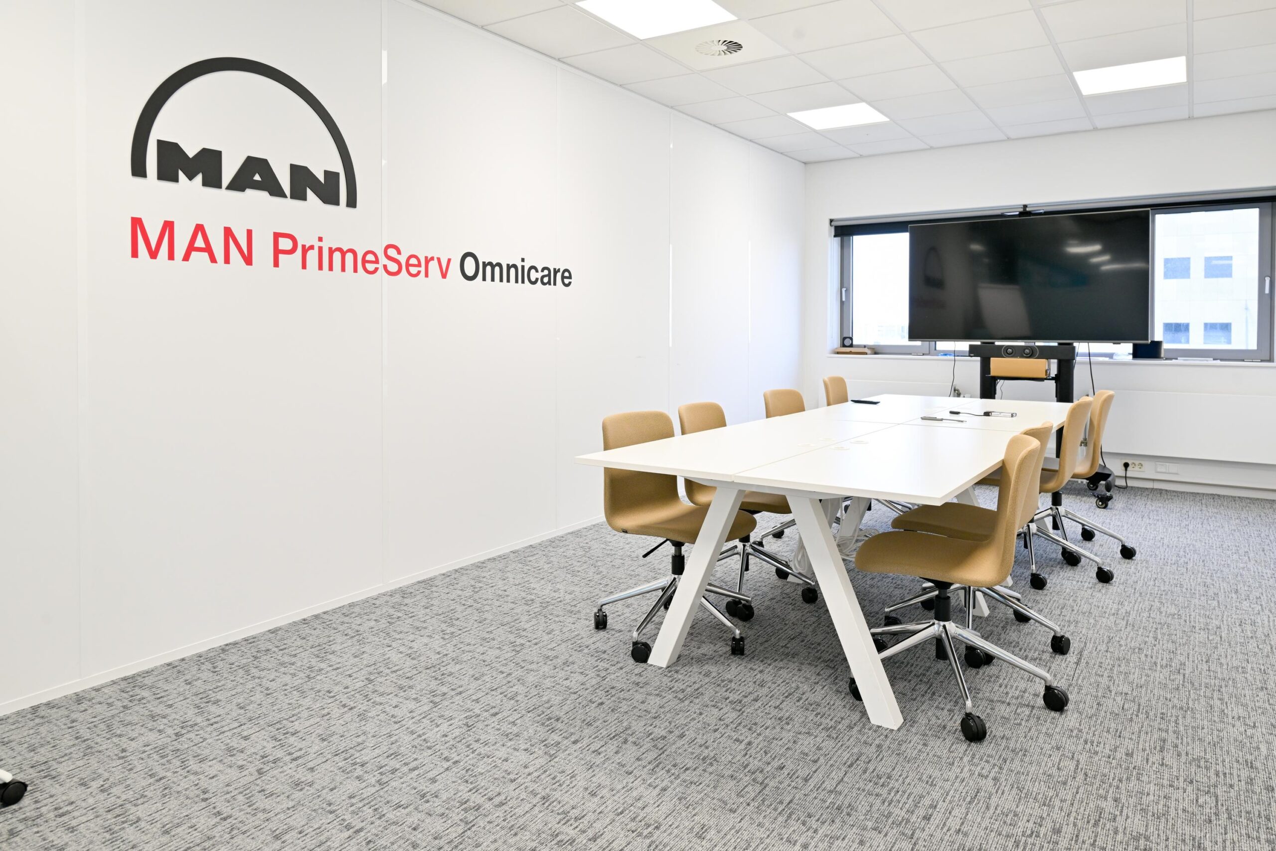 Vergaderruimte bij MAN PrimeServ Omnicare met witte tafels, beige stoelen en een groot scherm.