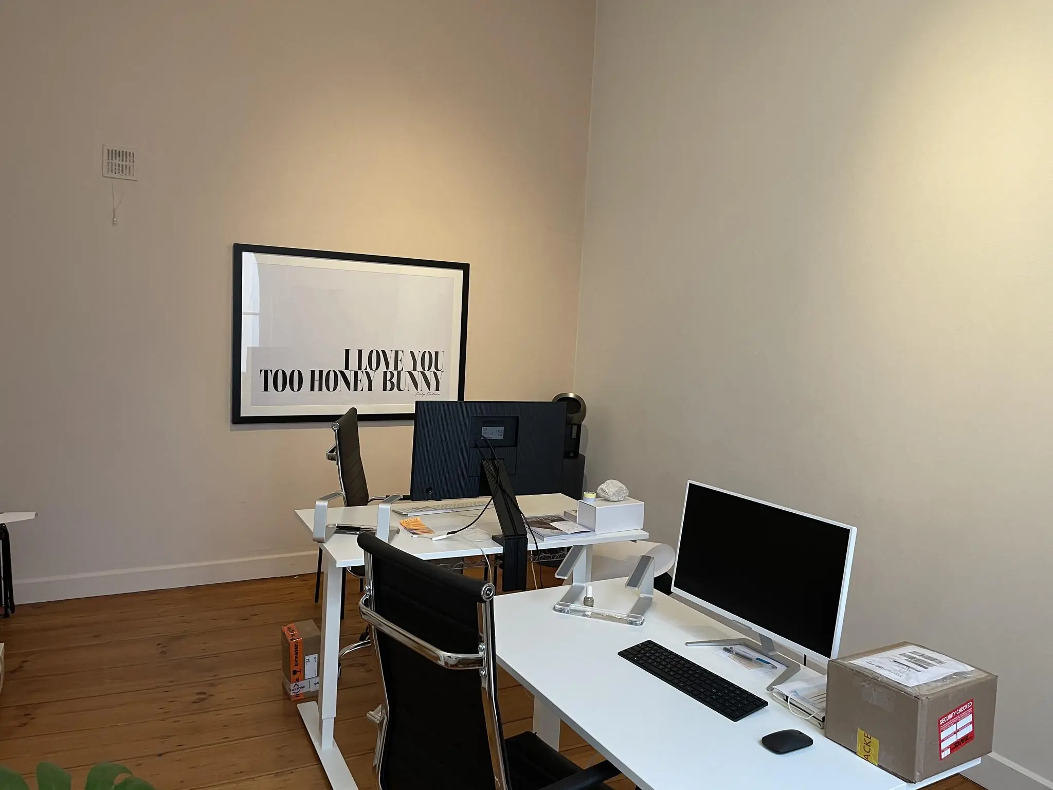 Moderne kantoorruimte met twee bureaus, computerschermen en een kunstwerk aan de muur met de tekst "I LOVE YOU TOO HONEY BUNNY".