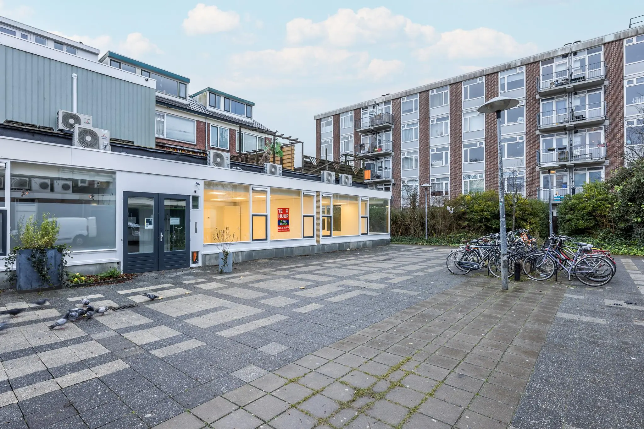 Winkelpand aan de Wallesteinlaan met 'Te Huur'-bord, omringd door appartementen, fietsenrek en geplaveid plein.