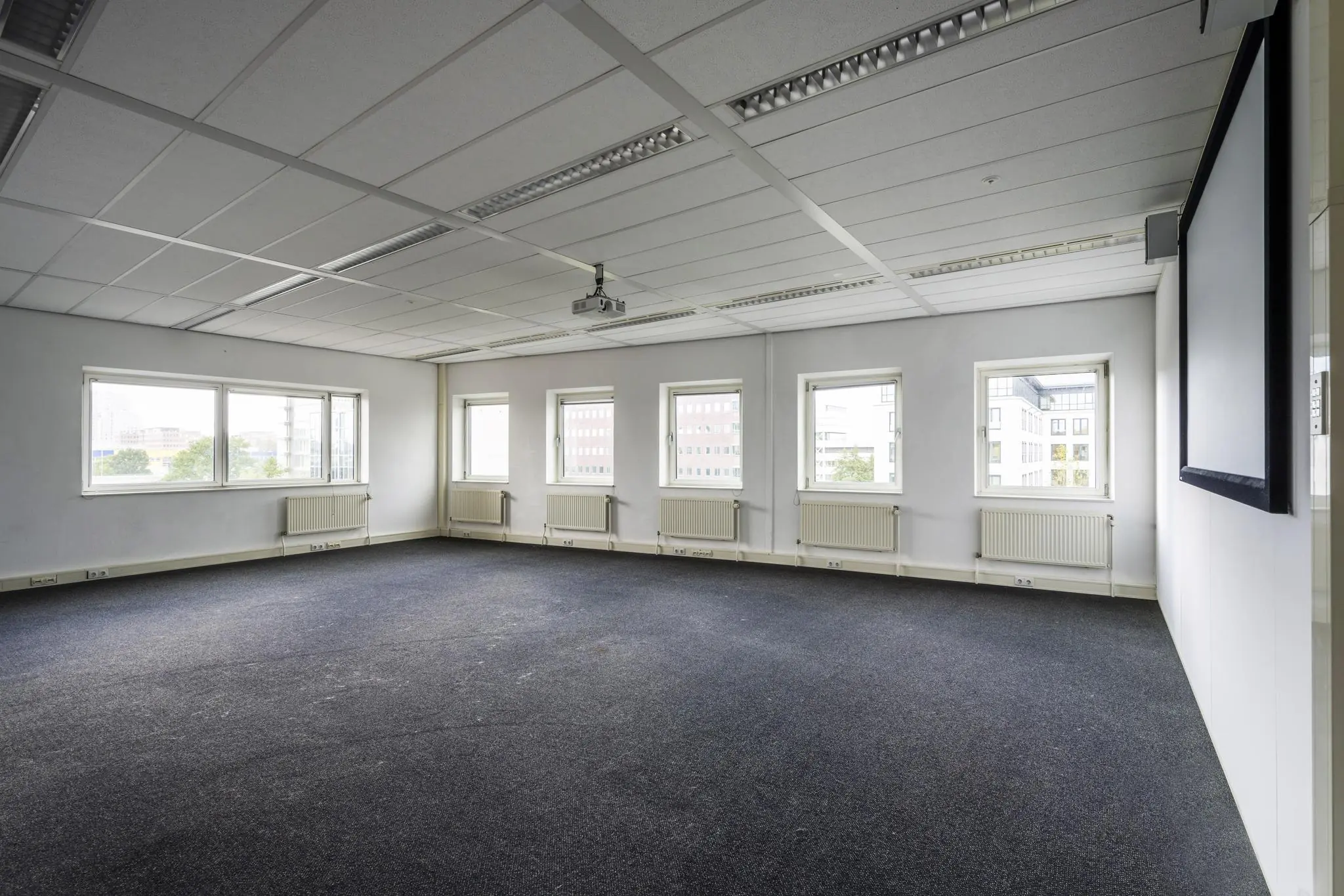 Leeg kantoor- of vergaderruimte aan de Paasheuvelweg met meerdere ramen, tapijt, projector en projectiescherm.