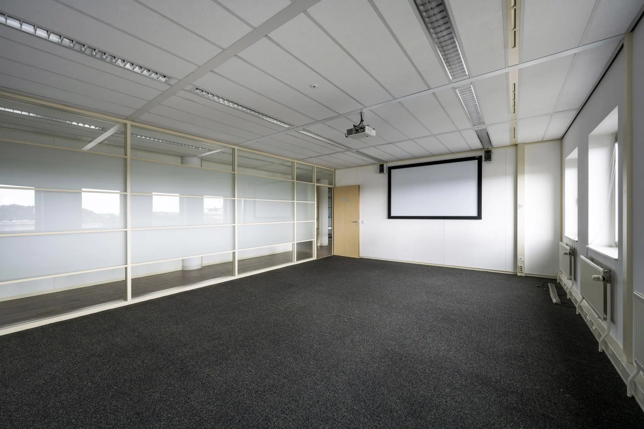 Lege vergaderruimte aan de Paasheuvelweg met projectiescherm, glazen scheidingswand en tapijtvloer.