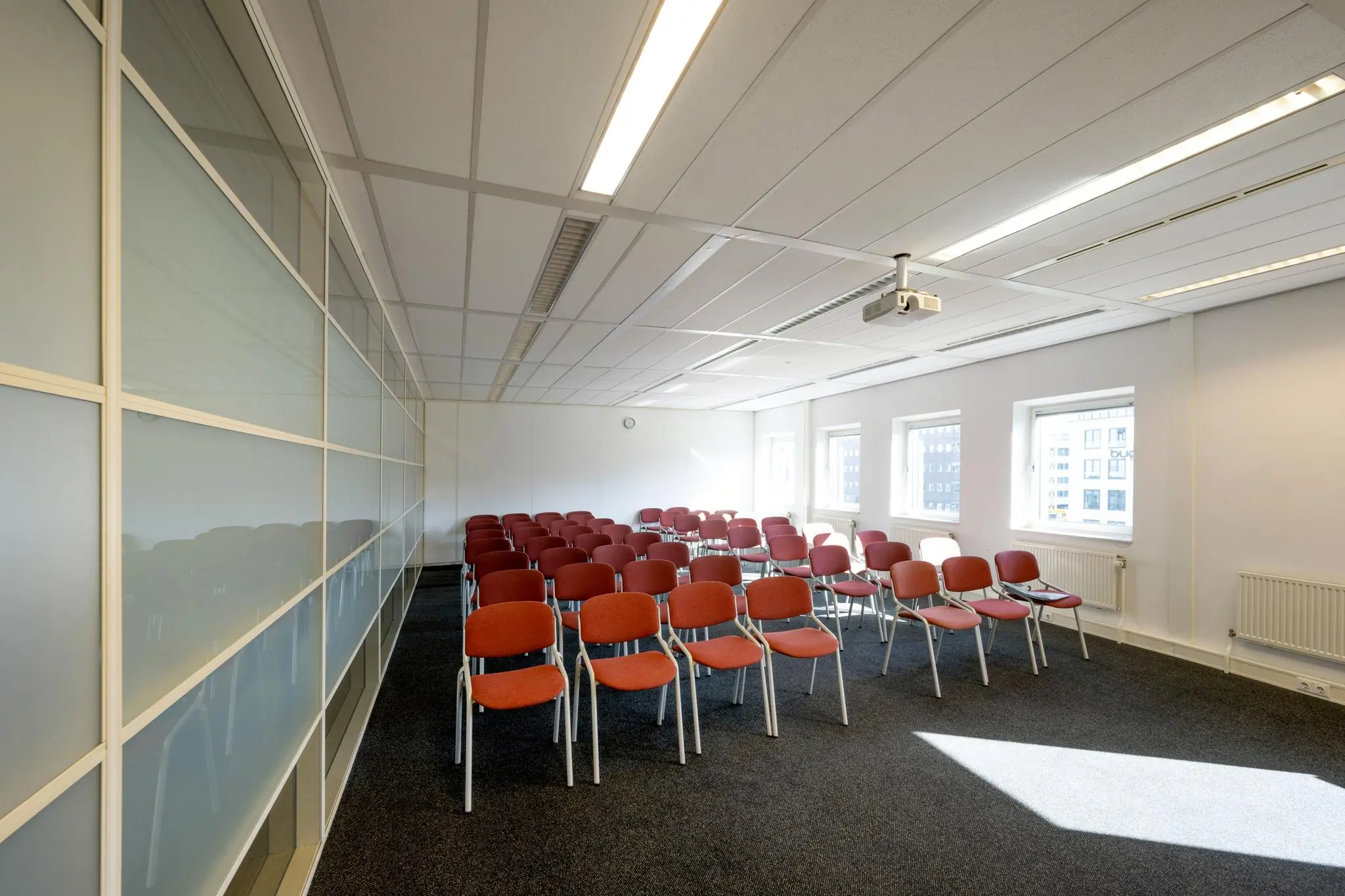 Vergaderruimte op de Paasheuvelweg met rijen rode stoelen, een projector aan het plafond en veel daglicht.