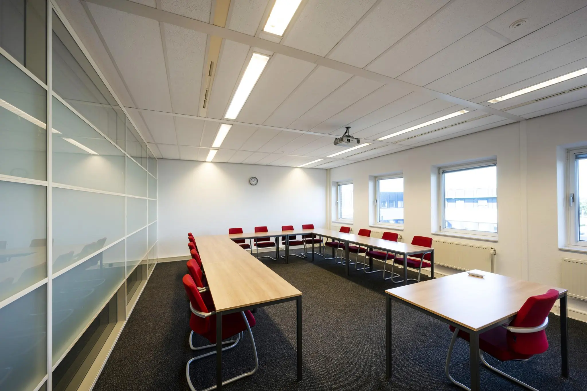 Vergaderruimte op de Paasheuvelweg met U-vormig opgestelde tafels, rode stoelen en een projector aan het plafond.