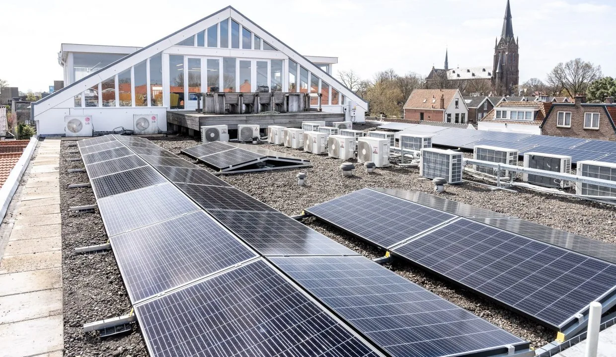 Zonnepanelen en airco-units op het platte dak van een gebouw aan het Oosteinde, met uitzicht op een kerk en woonhuizen.