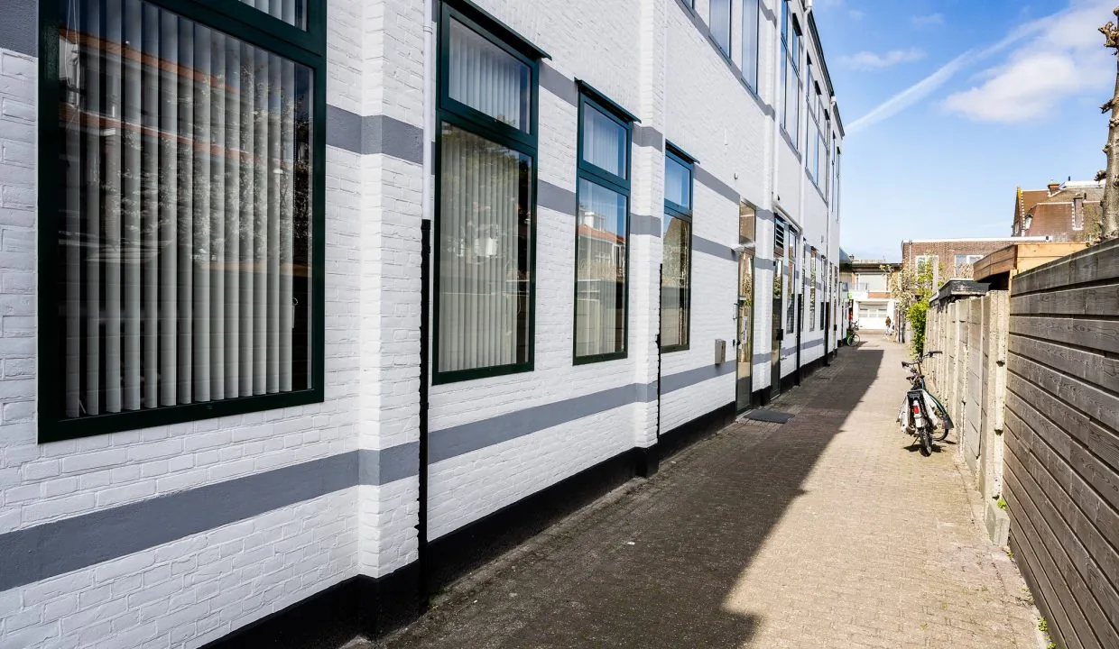 Smal steegje naast een wit gebouw aan het Oosteinde, met gestalde fiets en zonlicht op de gevels.