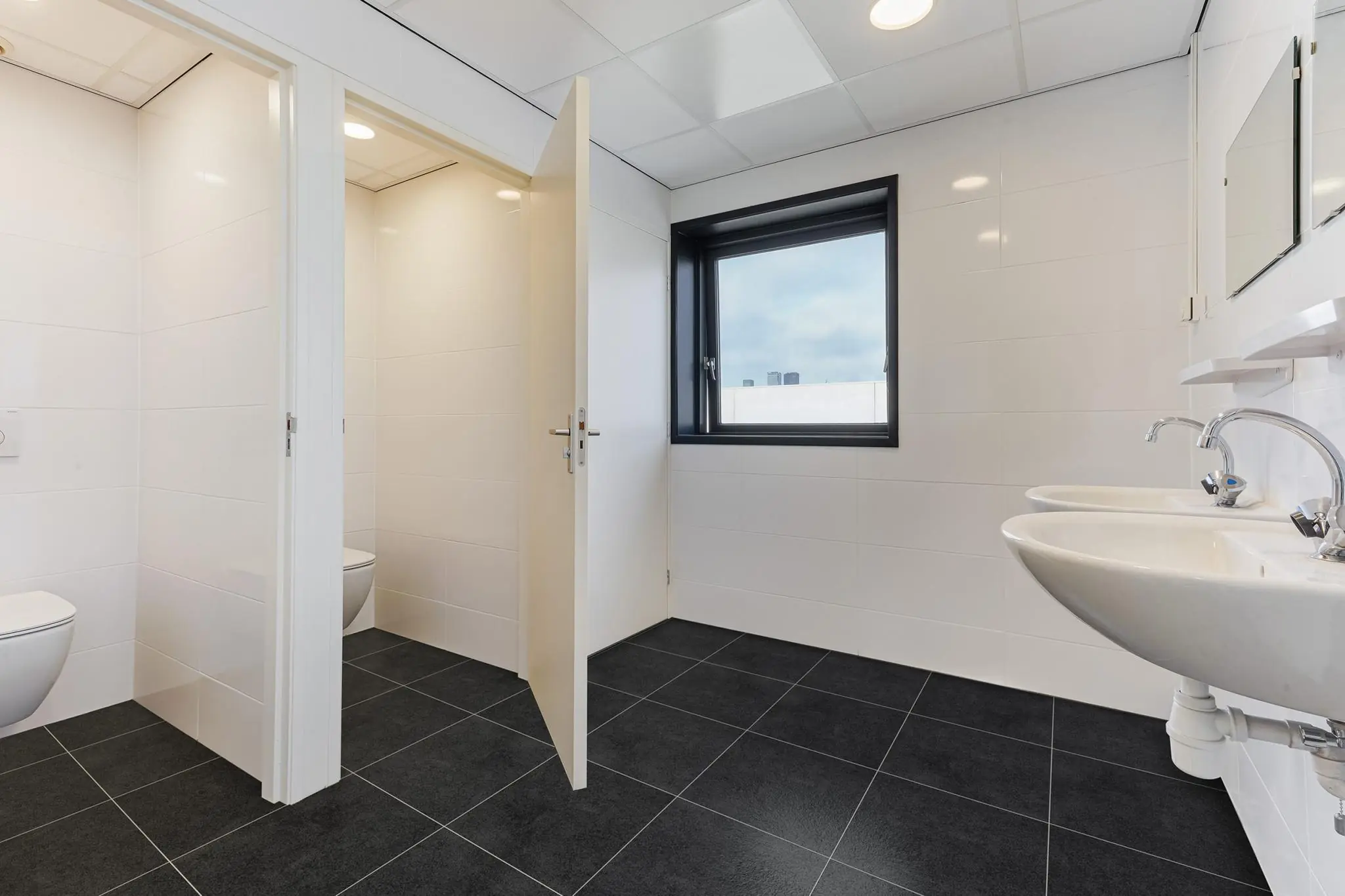Moderne sanitaire ruimte met twee toiletcabines, twee wastafels en een raam met daglicht.