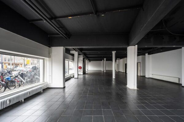Leegstaand zwart-wit interieur van een ruime commerciële ruimte aan de Muiderstraat met grote ramen en zicht op geparkeerde fietsen buiten.