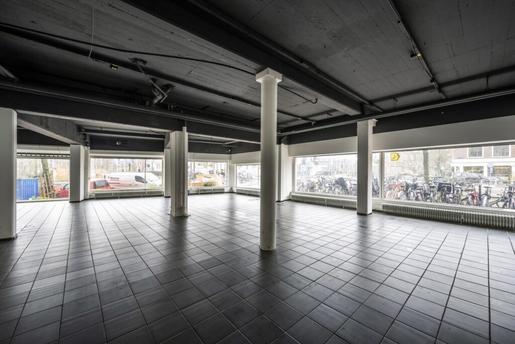 Leeg bedrijfsruimte aan de Muiderstraat met grote ramen en zwart betegelde vloer.