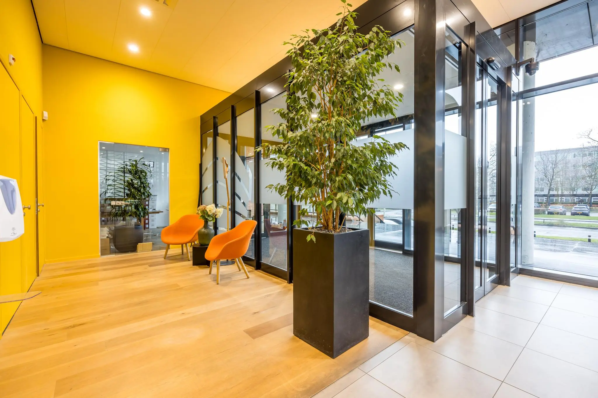 Moderne entreehal aan de Winthontlaan met grote raampartijen, felgele muur, planten en oranje stoelen.