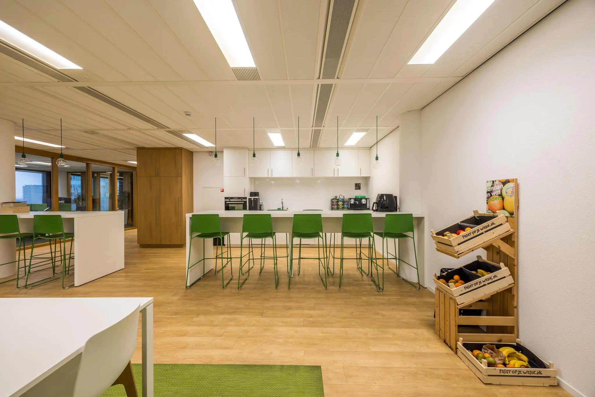 Moderne kantoorkeuken met een bar met groene barkrukken, hanglampen en een fruitrek van "Fruit op je werk".
