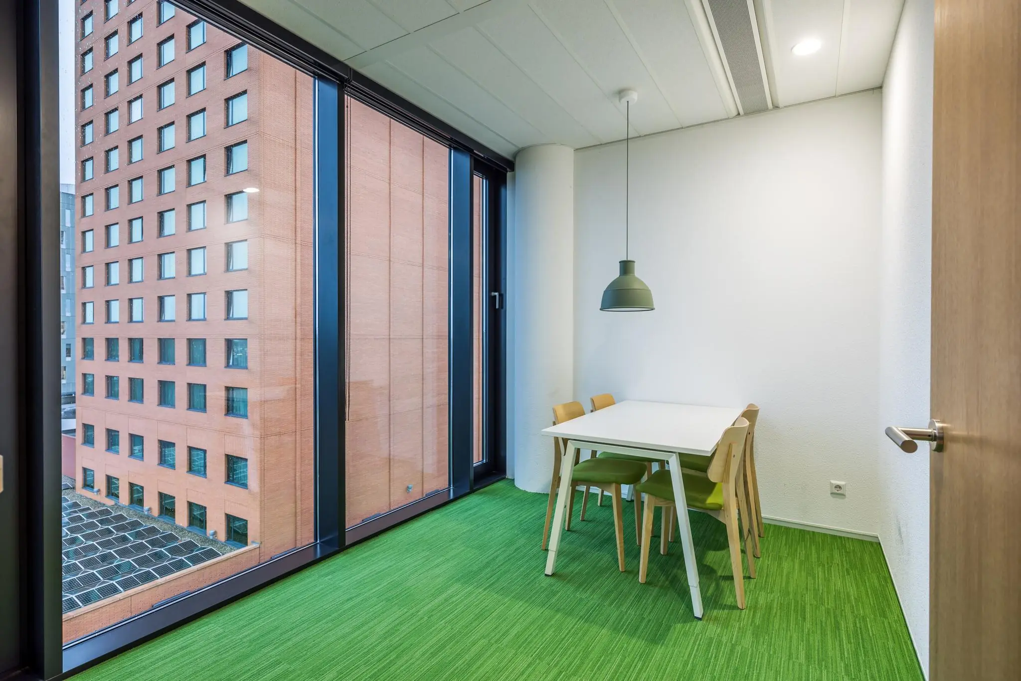 Kleine vergaderruimte aan de Winthontlaan met een witte tafel, vier stoelen en uitzicht op een rood bakstenen kantoorgebouw.