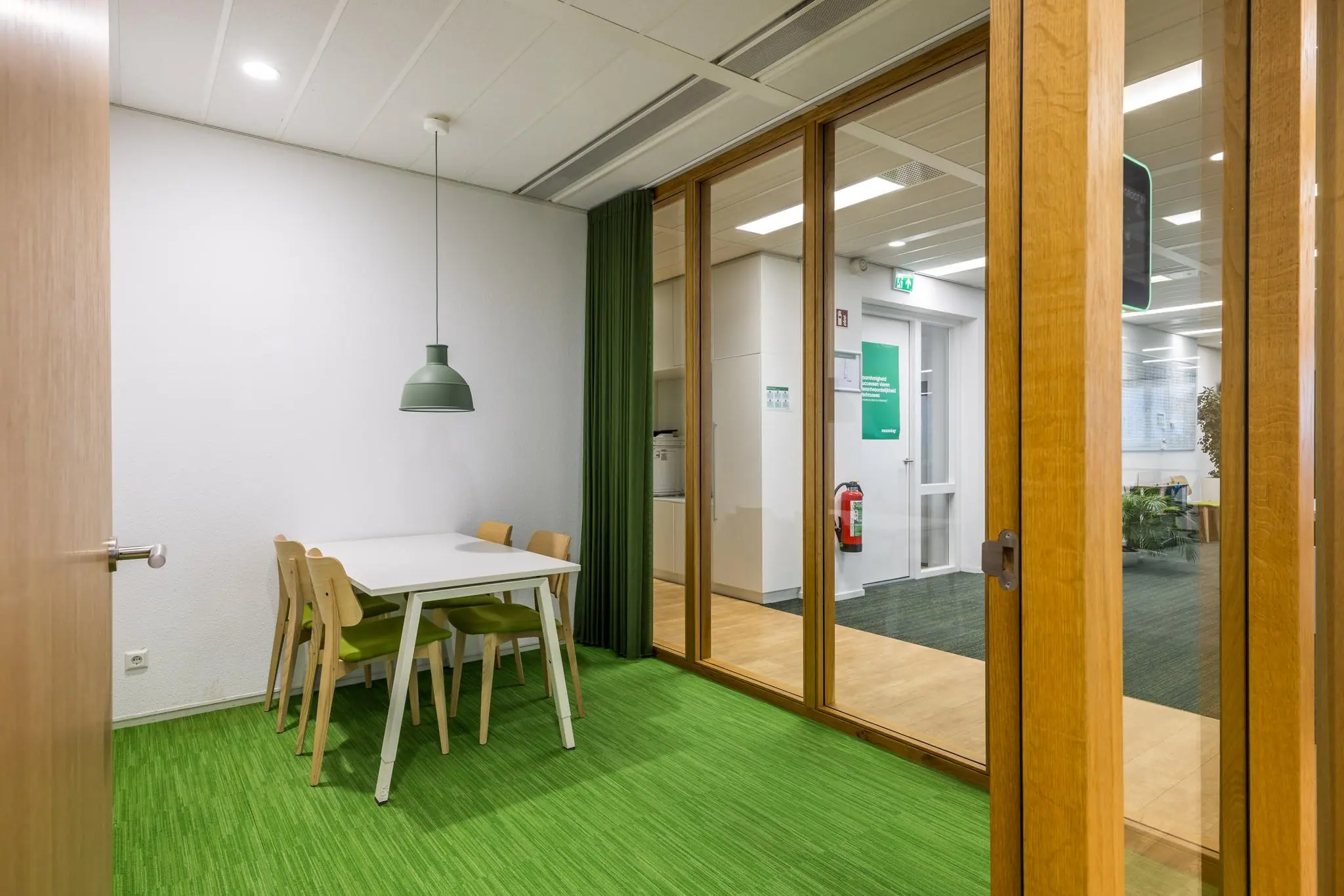 Vergaderruimte aan de Winthontlaan met witte tafel, groene stoelen, groene hanglamp en glazen wand met uitzicht op kantoorruimte.