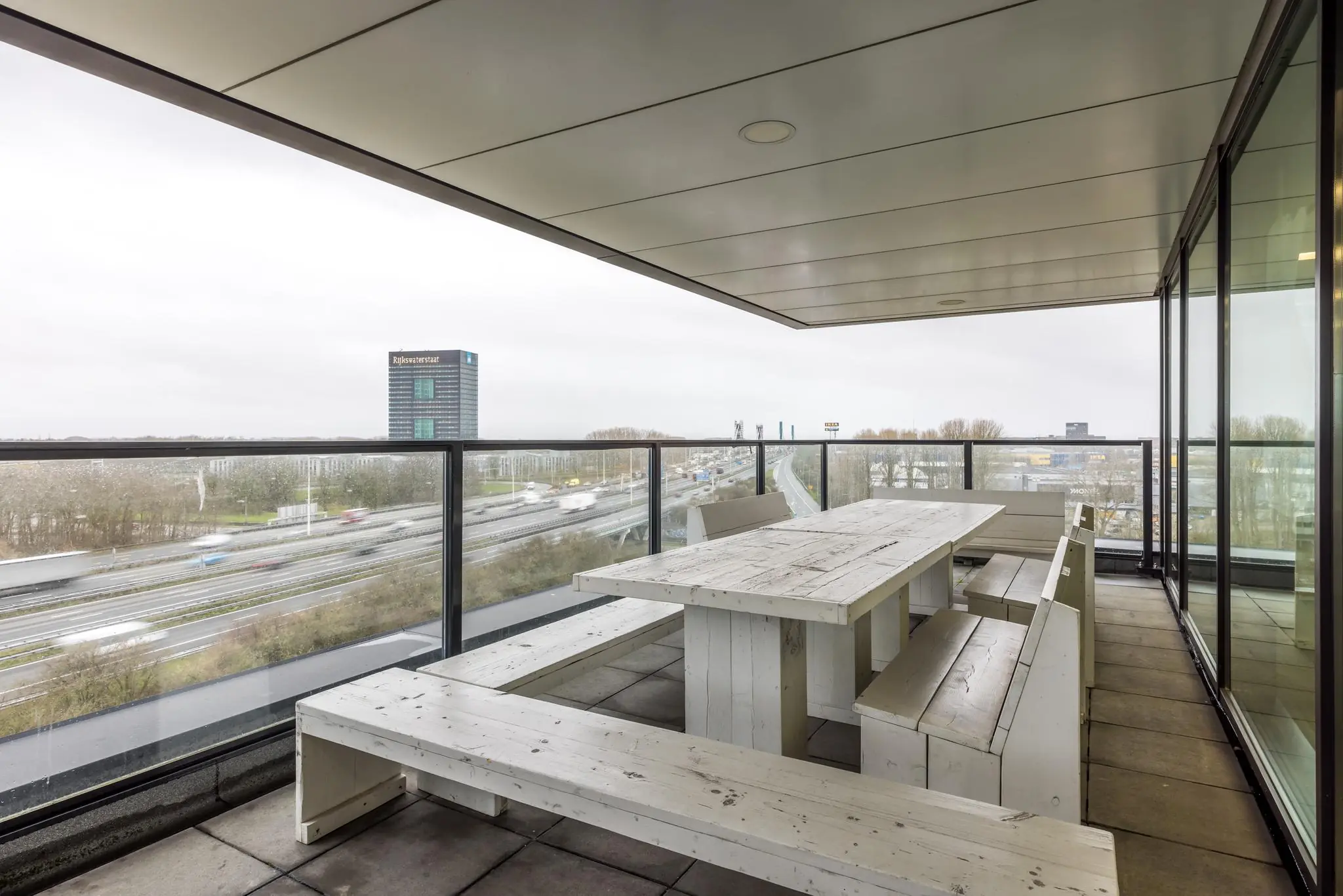 Overdekt balkon met houten picknicktafel en uitzicht op de snelweg en het Rijkswaterstaat-gebouw aan de Winthontlaan.