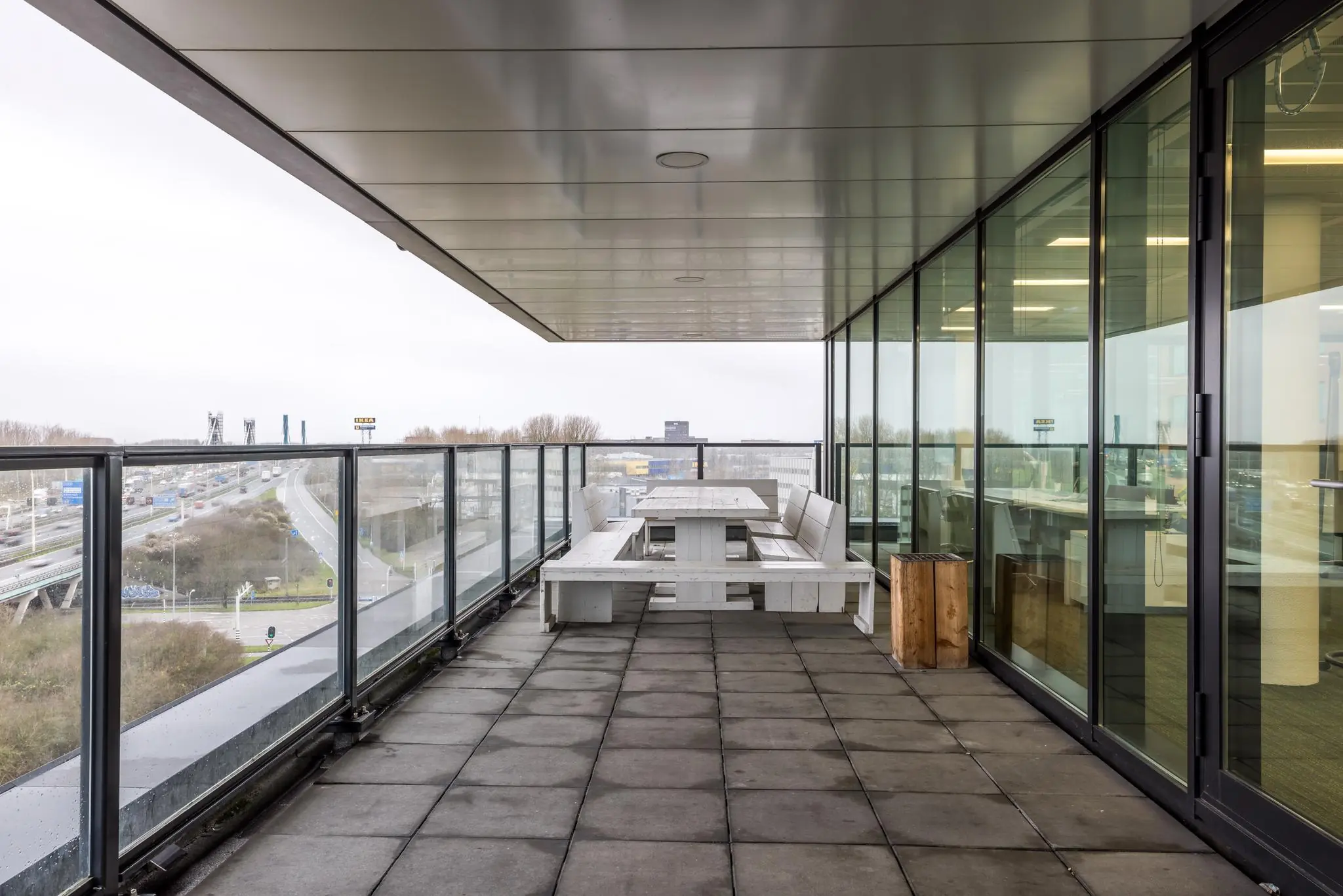 Overdekt kantoorbalkon aan de Winthontlaan met glazen balustrade, witte picknicktafels en uitzicht op snelweg en stad.
