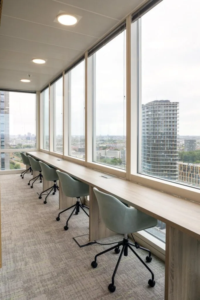 Modern ingerichte flexwerkplekken langs een raam met uitzicht op Amstelplein en een hoog kantoorgebouw.