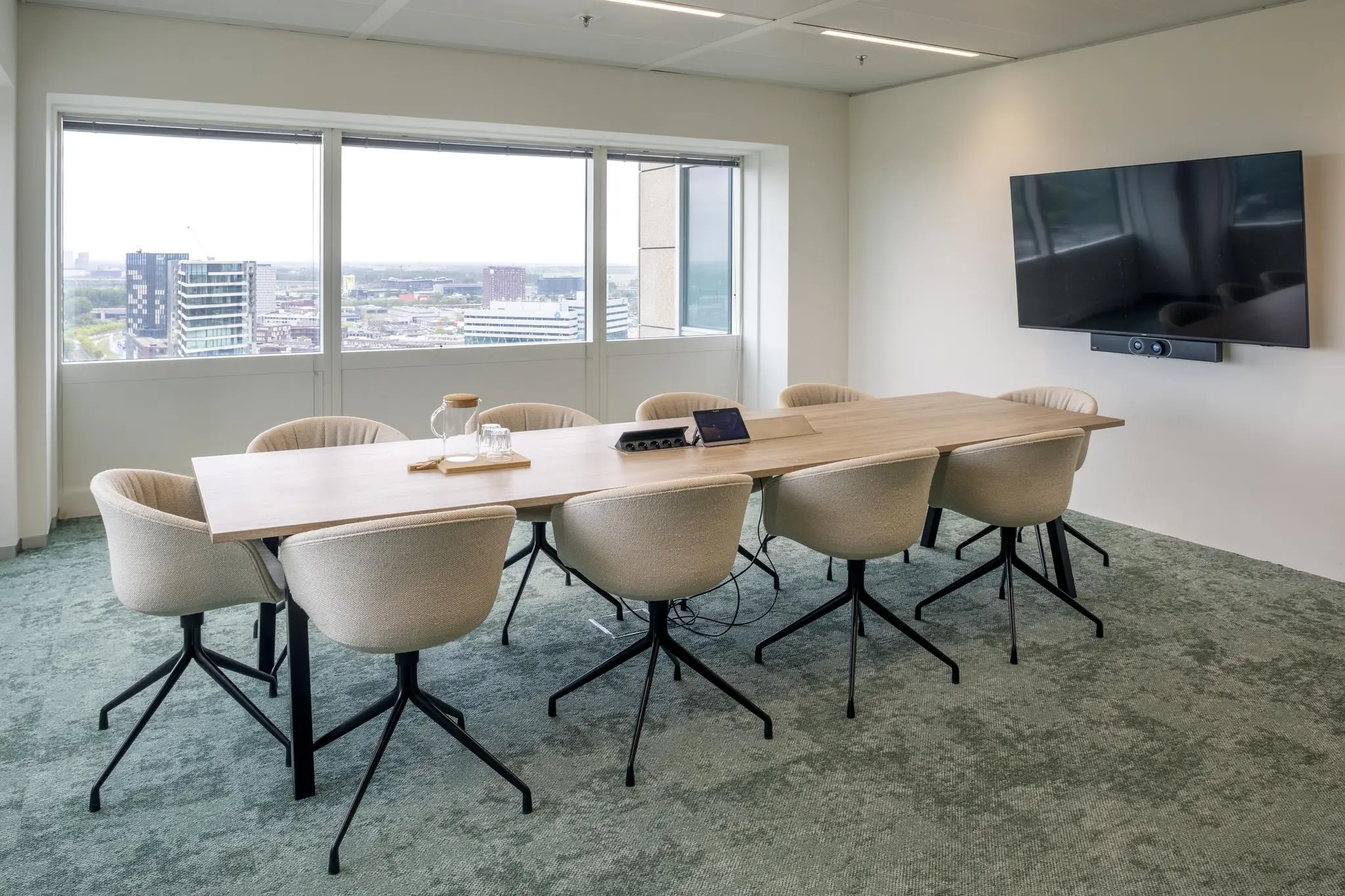 Vergaderkamer in Amstelplein met een lange houten tafel, acht stoelen, een flatscreen en uitzicht op moderne kantoorgebouwen.