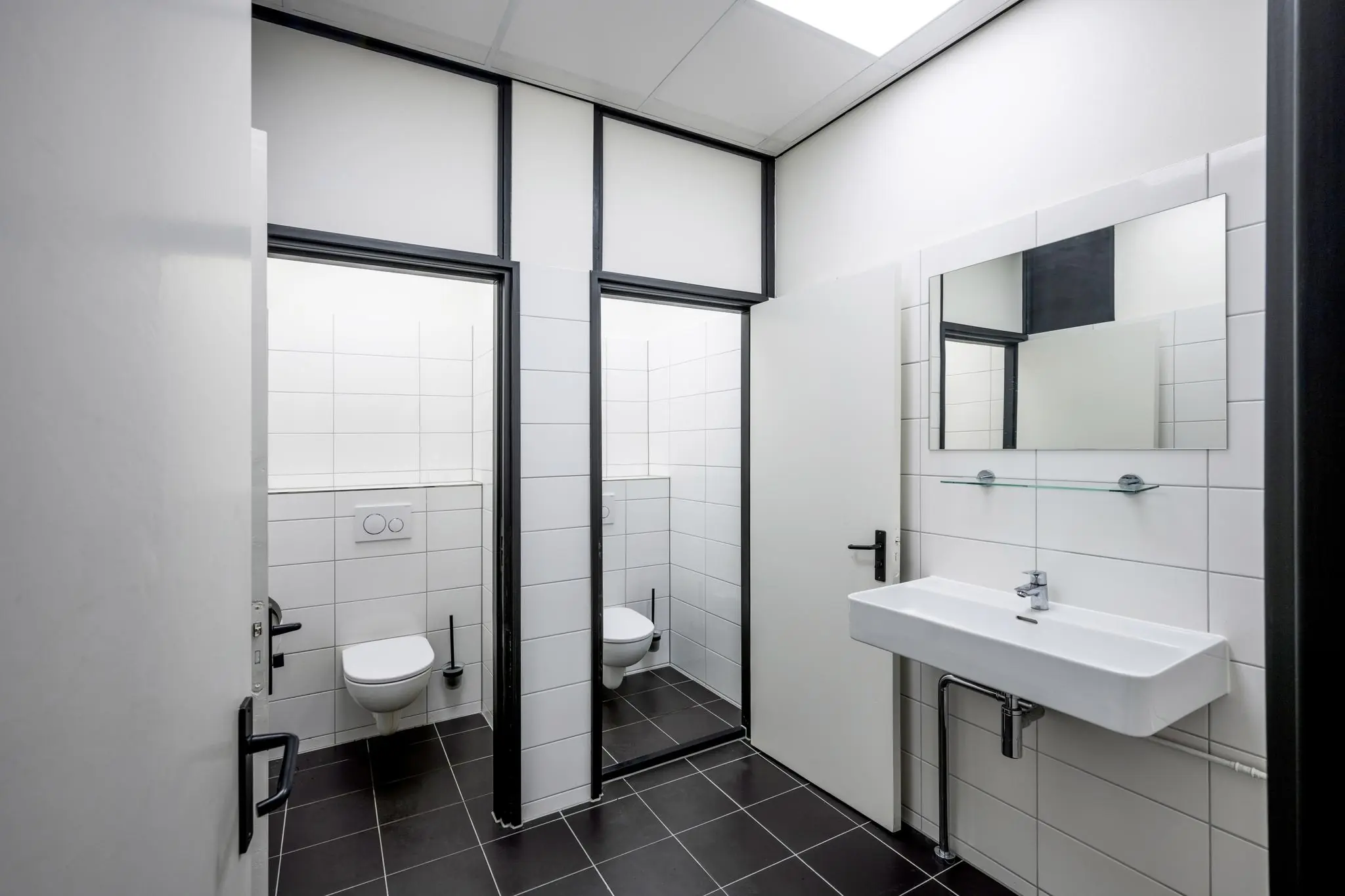 Moderne, wit betegelde gedeelde toiletruimte met twee aparte wc-hokjes en een lange wastafel met spiegel.