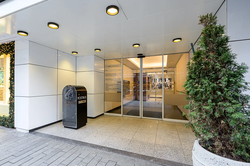 Ingang van het Minerva Huis aan de Meent met glazen schuifdeuren, een postbus en een grote plantenbak.