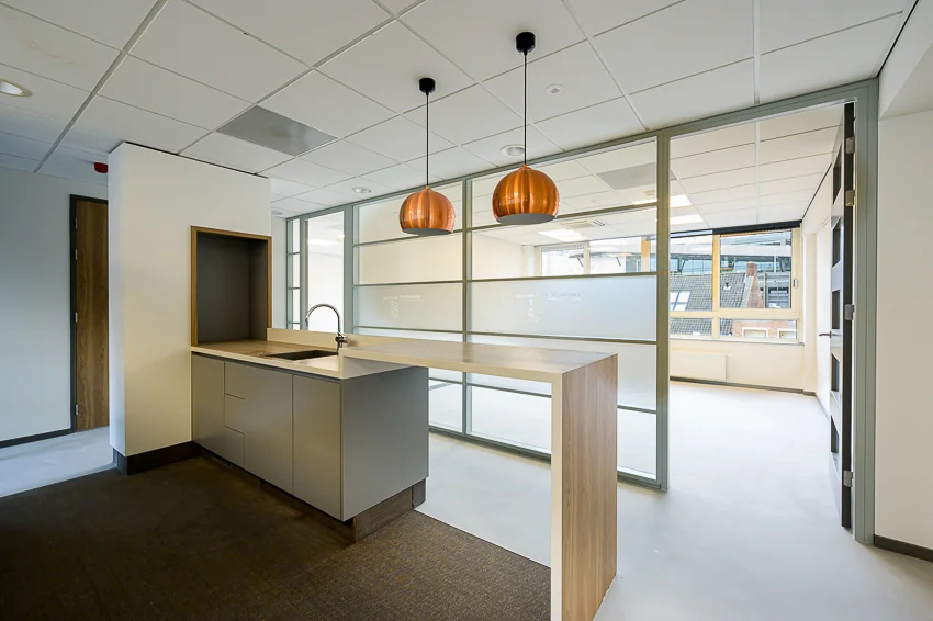 Moderne kantoorruimte met kitchenette, glazen scheidingswanden en hanglampen aan het plafond.