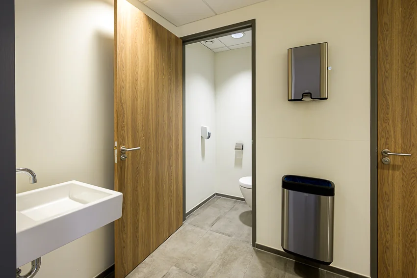 Moderne, schone toiletruimte met open deur naar wc, witte wastafel, prullenbak en papieren handdoekdispenser.