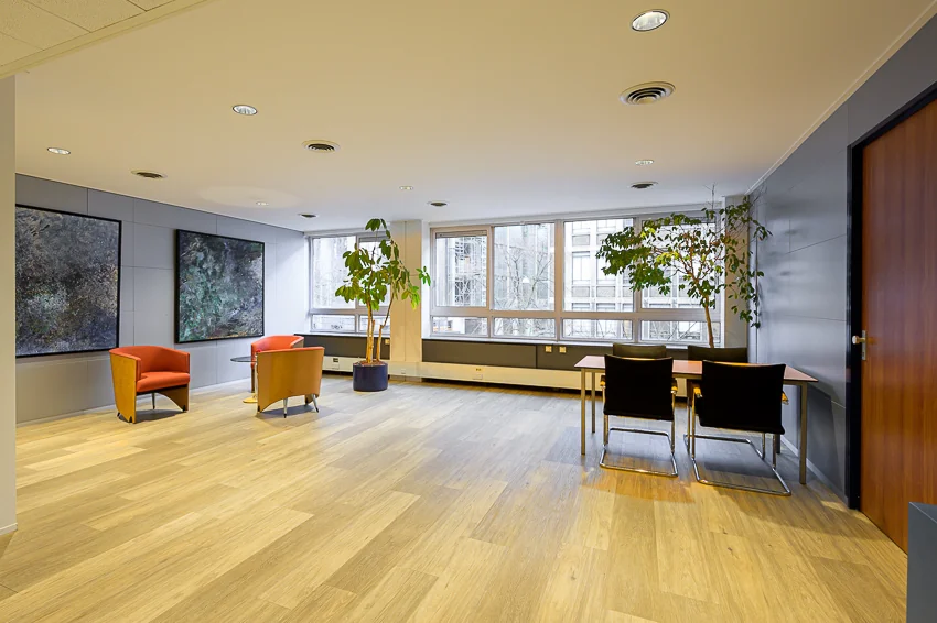 Moderne wacht- of vergaderruimte op de Meent, met houten vloer, grote ramen, kamerplanten en kleurige zitstoelen.