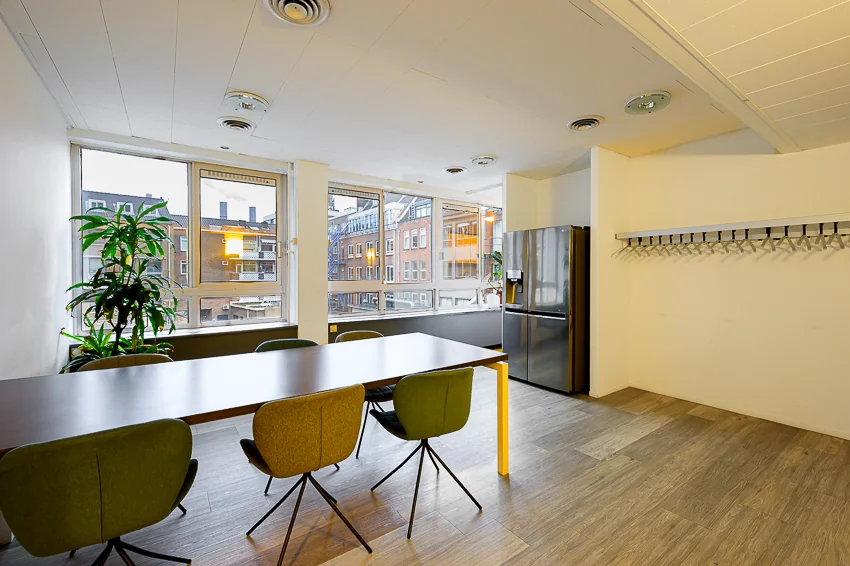 Moderne gezamenlijke ruimte met grote ramen, een lange tafel met stoelen, een roestvrijstalen koelkast en uitzicht op de Meent in Rotterdam.
