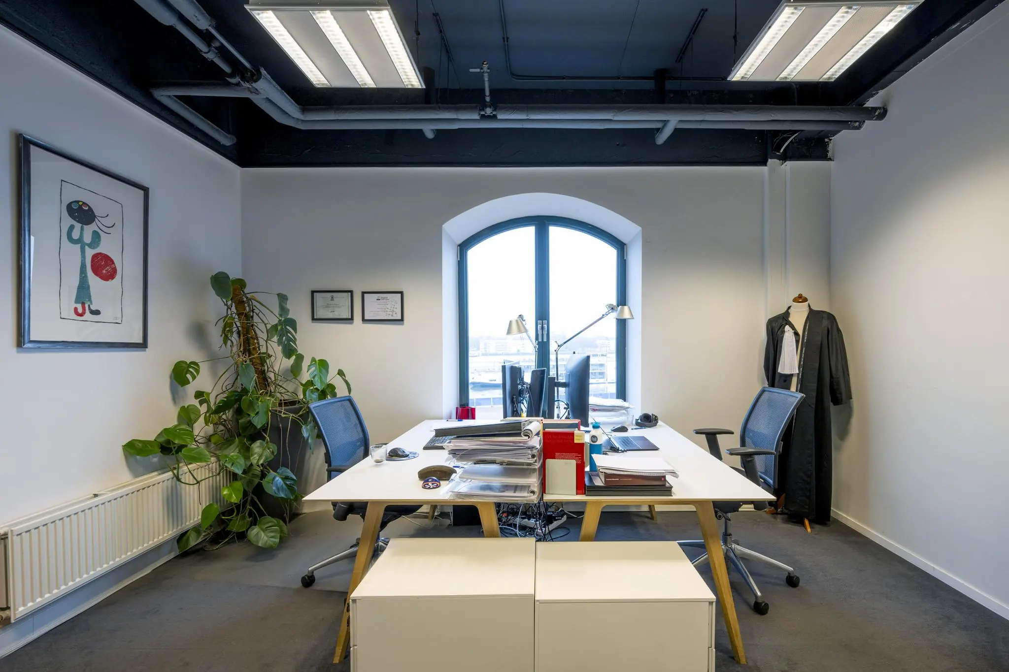 Modern kantoor aan de De Ruijterkade met twee bureaus, een toga op een paspop en uitzicht op de stad.