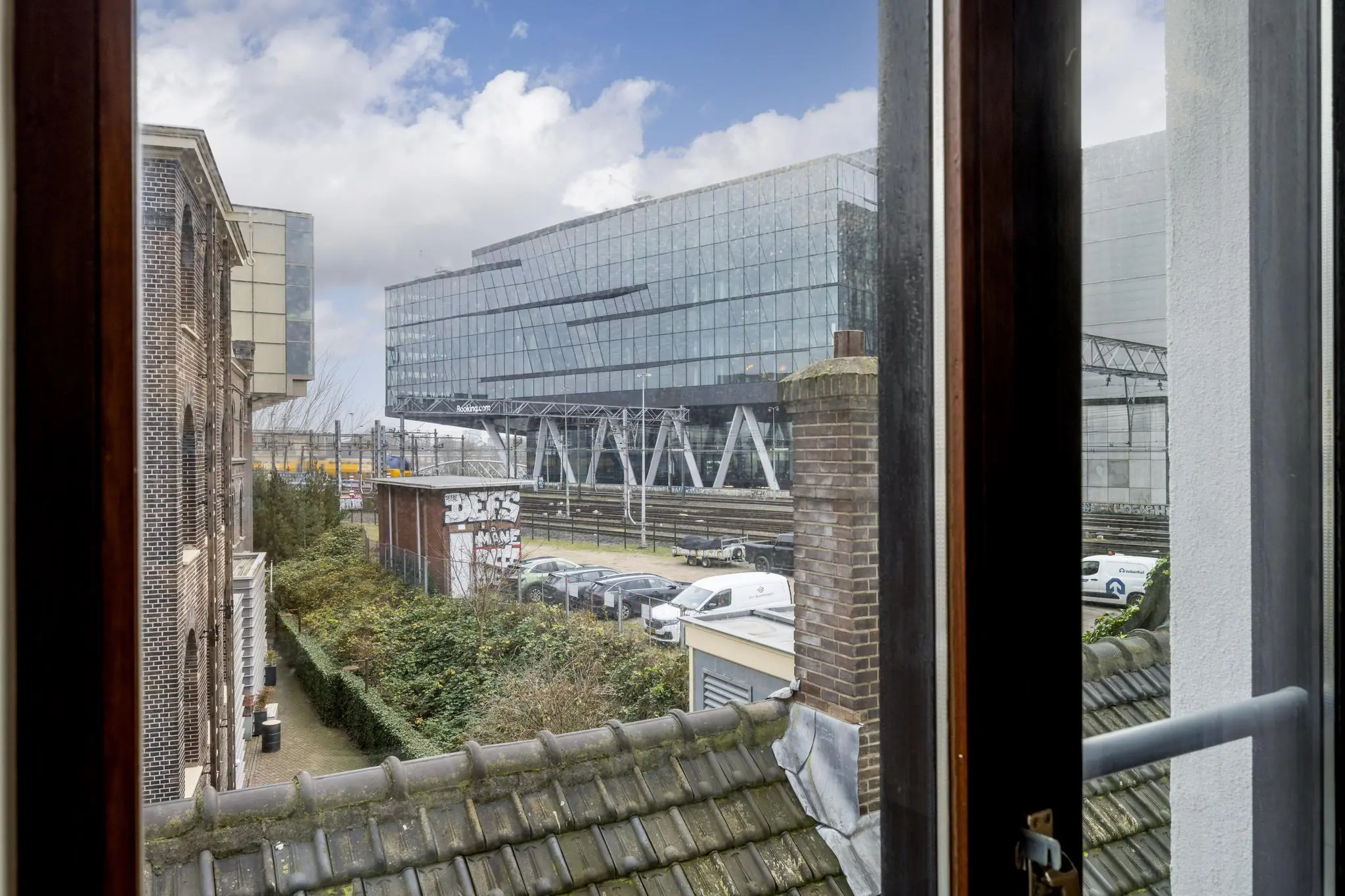 Uitzicht vanaf een raam op De Ruijterkade met moderne kantoorgebouwen, spoorlijnen en geparkeerde auto's.