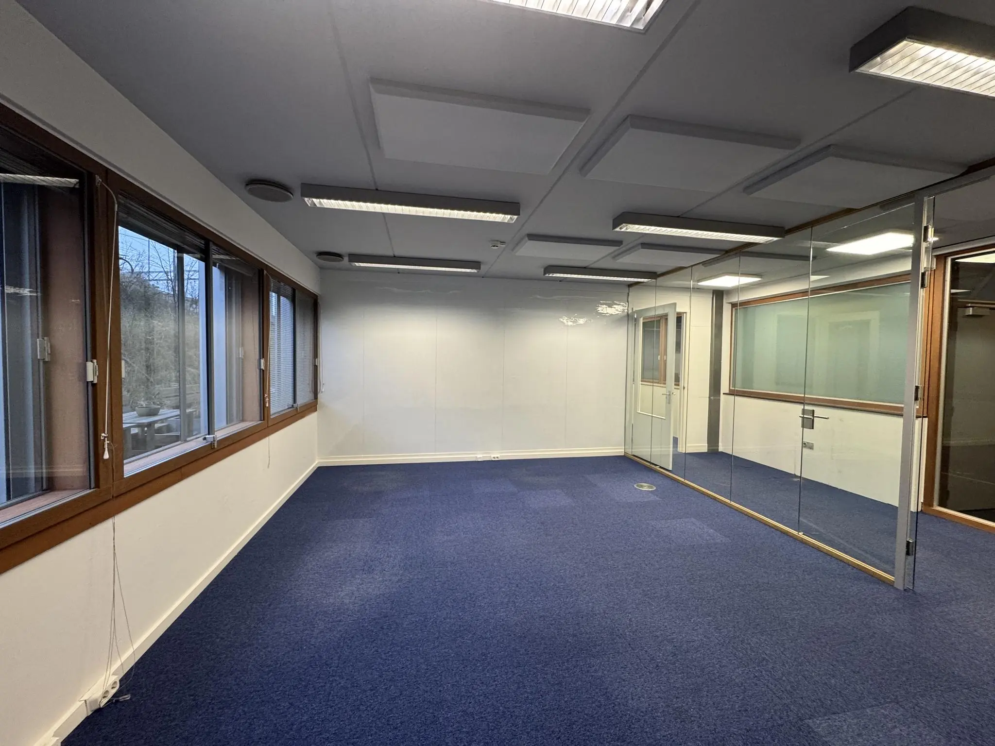 Leeg kantoor aan de De Ruijterkade met blauwe vloerbedekking, grote ramen en een glazen scheidingswand.