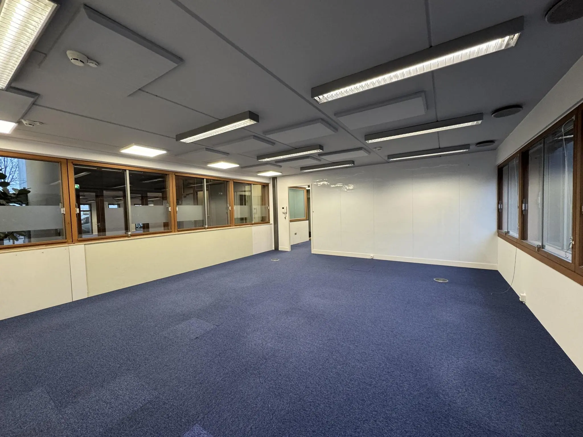 Leeg kantoorpand aan de De Ruijterkade met blauw tapijt, systeemplafond en grote ramen met houten kozijnen.