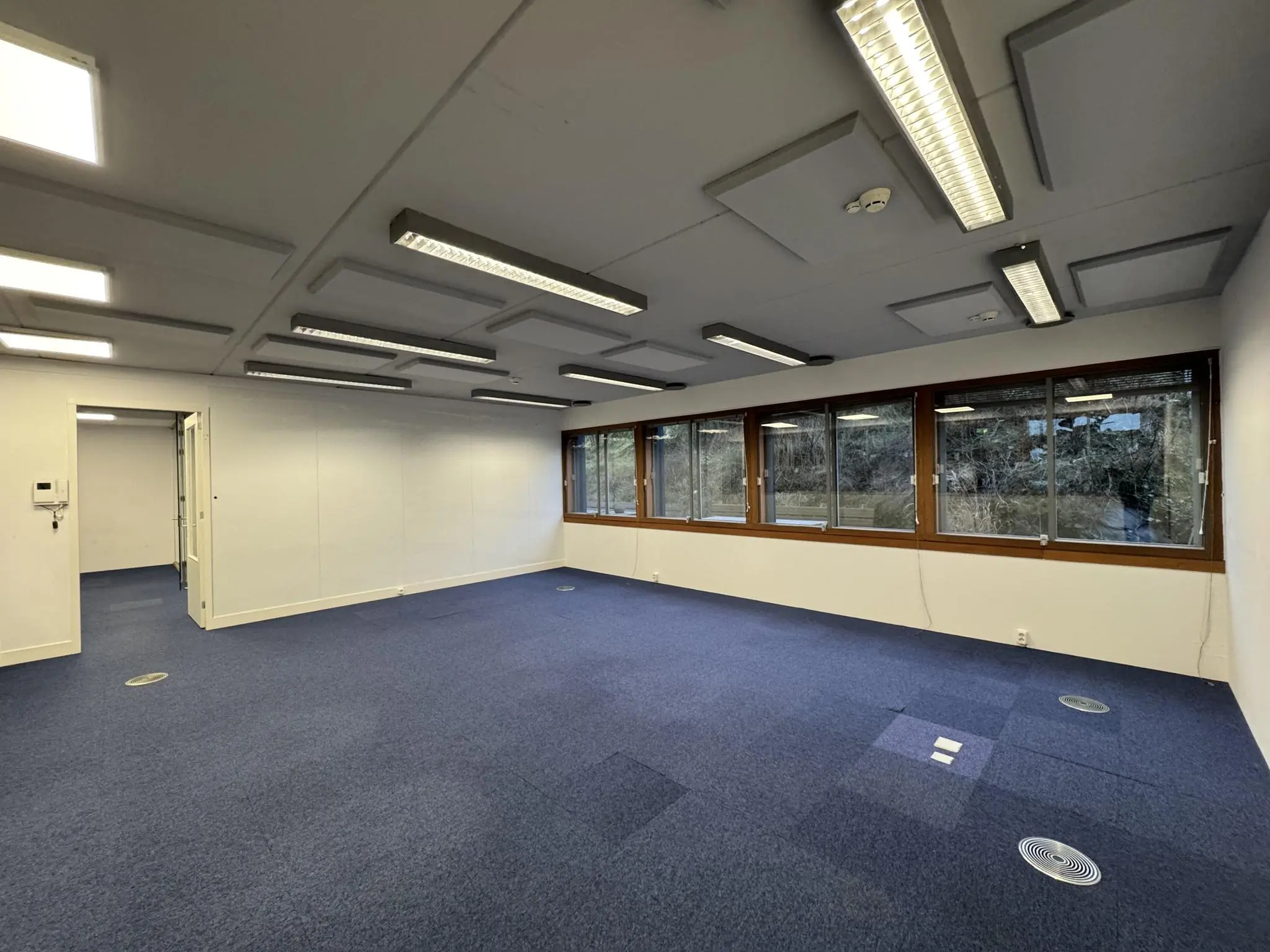 Lege kantoorruimte aan de De Ruijterkade met blauwe vloerbedekking, witte wanden en grote ramen met uitzicht op buiten.