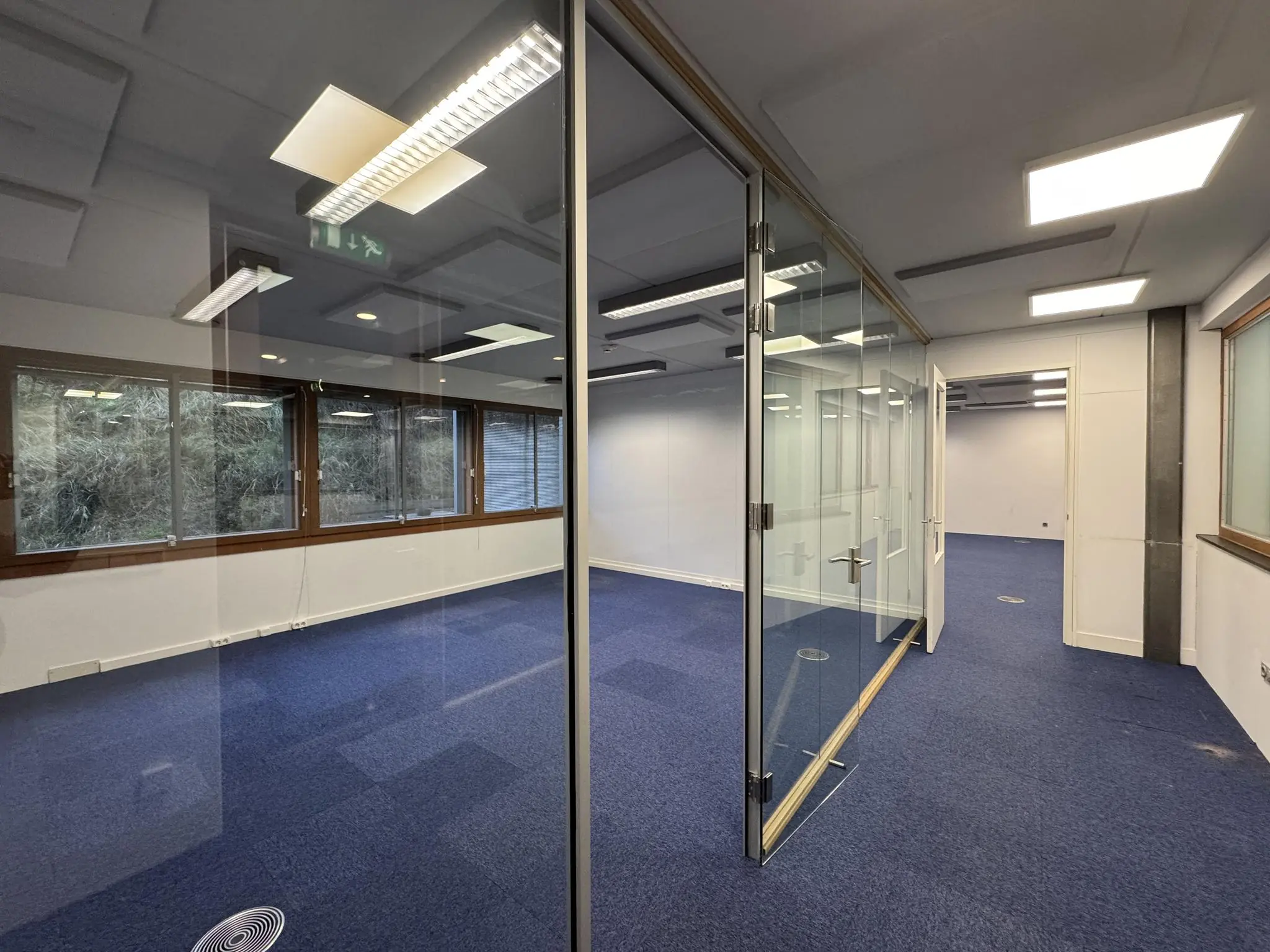 Kantoorruimte aan de De Ruijterkade met blauwe vloerbedekking, glazen scheidingswanden en grote ramen met uitzicht op groen.