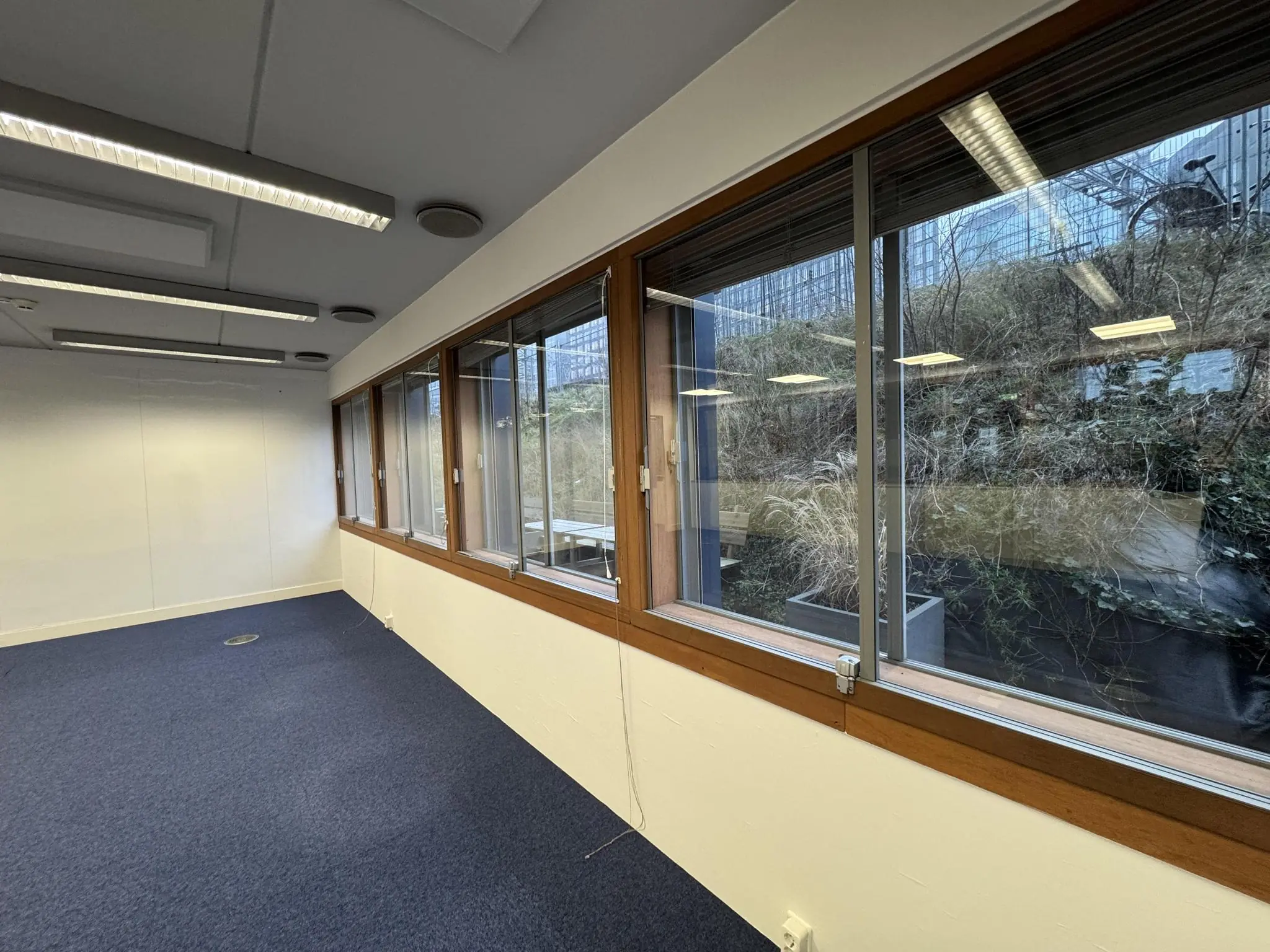 Lege kantoorruimte aan de De Ruijterkade met grote ramen en uitzicht op een verhoogde tuin en moderne gebouwen.