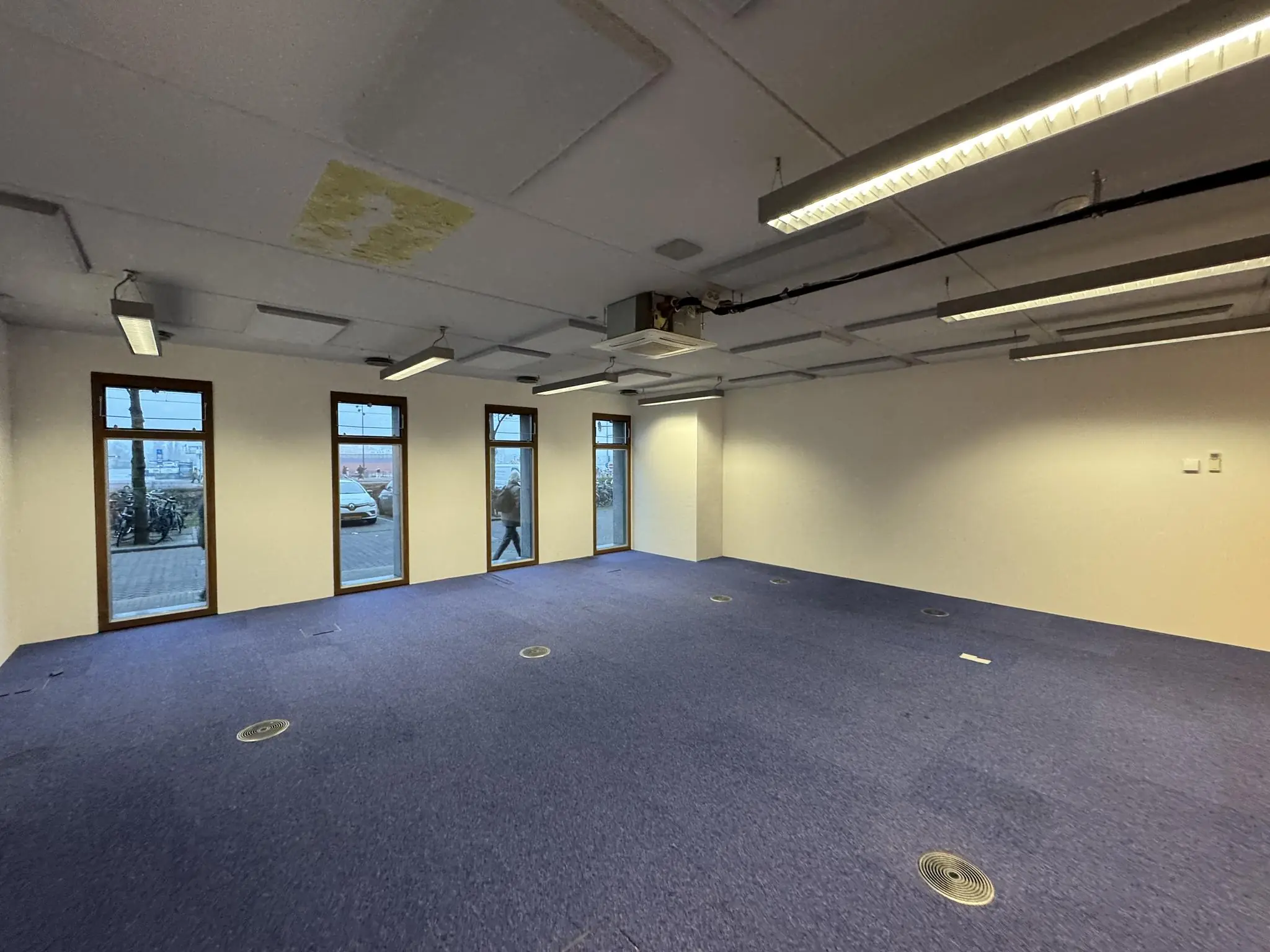 Leeg kantoorpand aan de De Ruijterkade met paarse vloerbedekking, witte muren en grote ramen met uitzicht op straat.