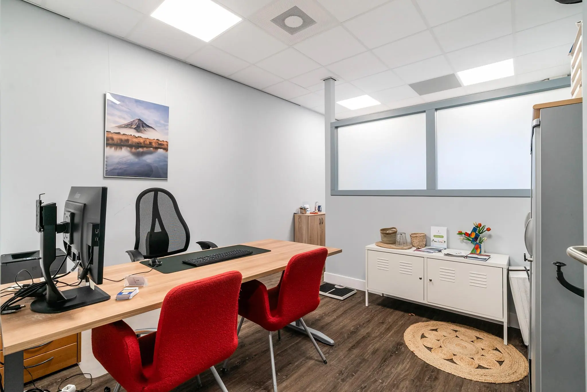 Modern ingerichte kantoorruimte aan de Bordewijklaan met twee rode stoelen, een houten bureau en een fotoprint van een berglandschap aan de muur.
