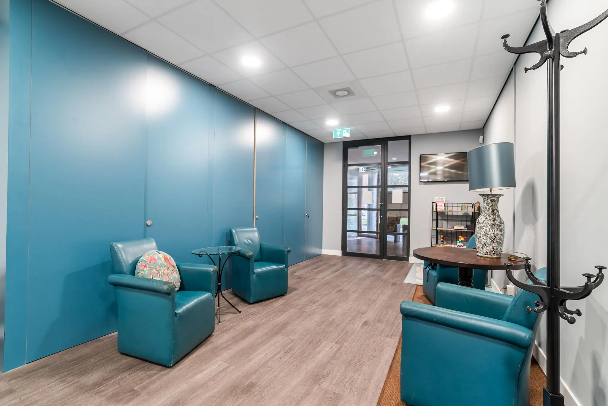 Wachtkamer met blauwe leren stoelen, een glazen tafeltje, kapstok en zicht op de entree aan de Bordewijklaan.