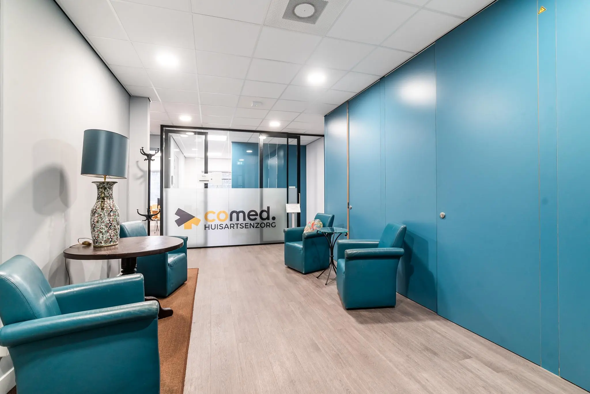 Wachtkamer van huisartsenpraktijk Comed met blauwe stoelen, een tafellamp en glazen deur met logo.