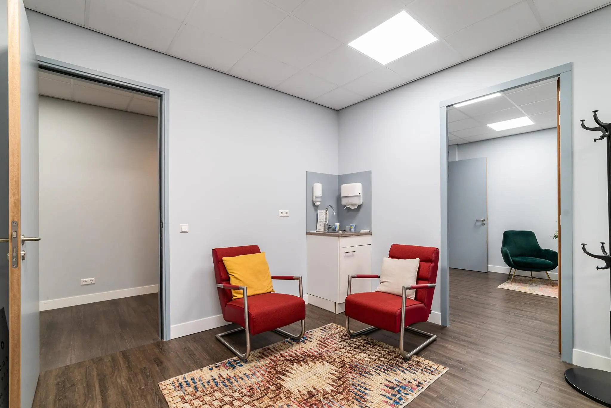 Wachtkamer met twee rode stoelen, een vloerkleed, een wastafel en zicht op een aangrenzende ruimte met een groene stoel.