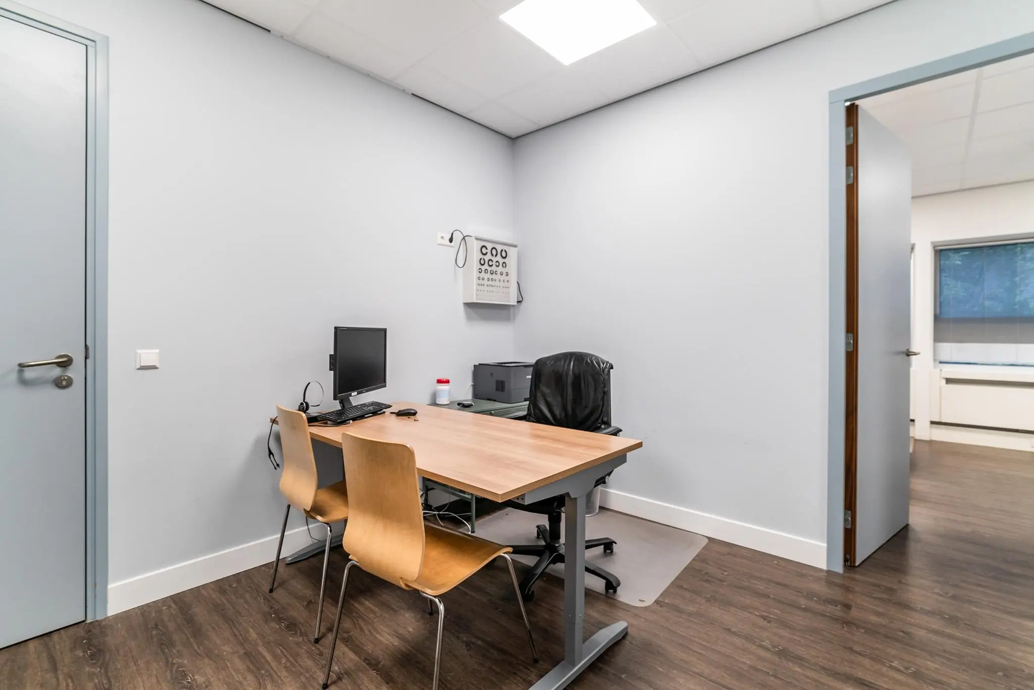 Kleine, moderne spreekkamer met bureau, computer en oogtestkaart aan de muur in een praktijk aan de Bordewijklaan.