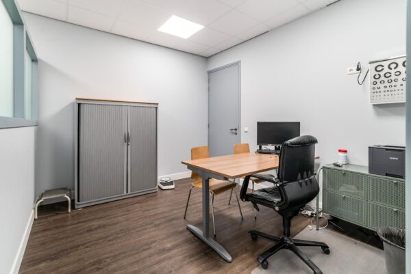 Kleine spreekkamer of dokterskantoor aan de Bordewijklaan met bureau, stoelen, computer en oogtestkaart aan de muur.