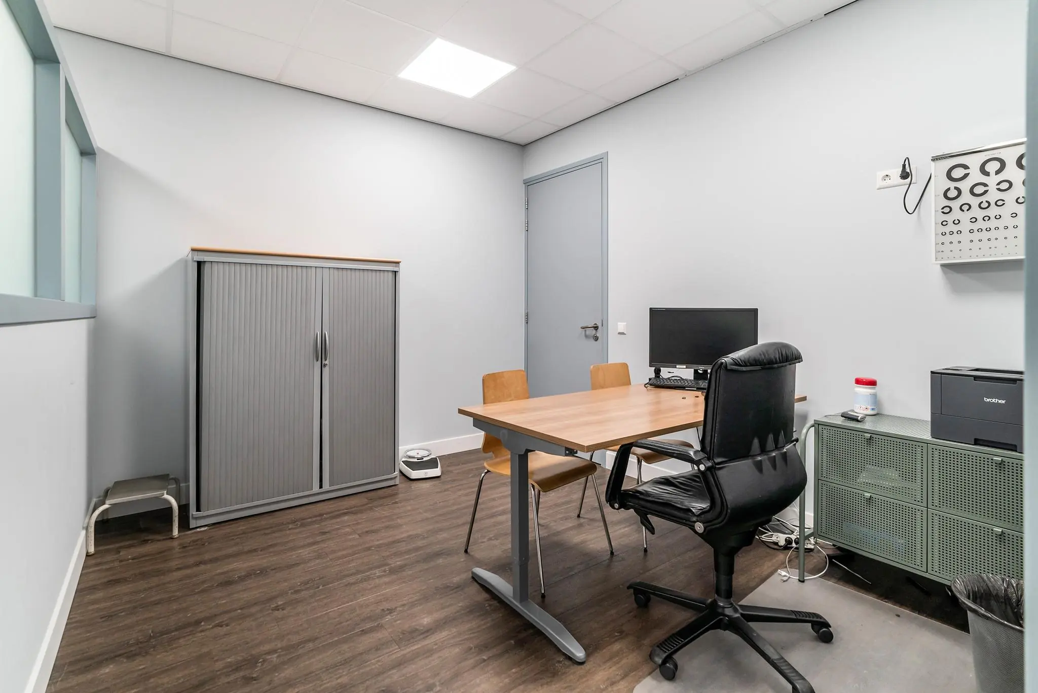 Kleine spreekkamer of dokterskantoor aan de Bordewijklaan met bureau, stoelen, computer en oogtestkaart aan de muur.