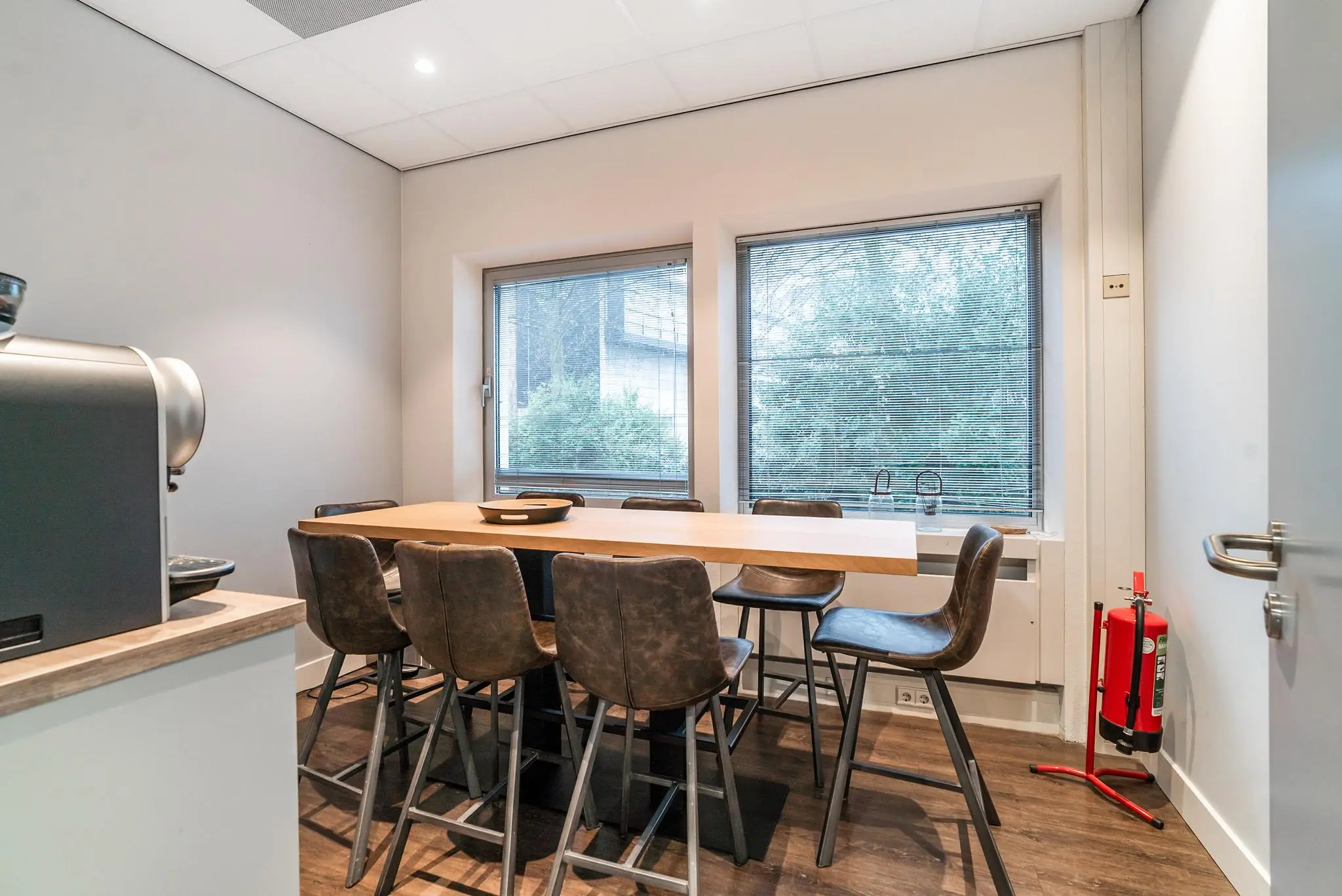 Vergaderruimte aan de Bordewijklaan met hoge tafel, leren barkrukken, koffiemachine en uitzicht op groen door twee ramen met jaloezieën.