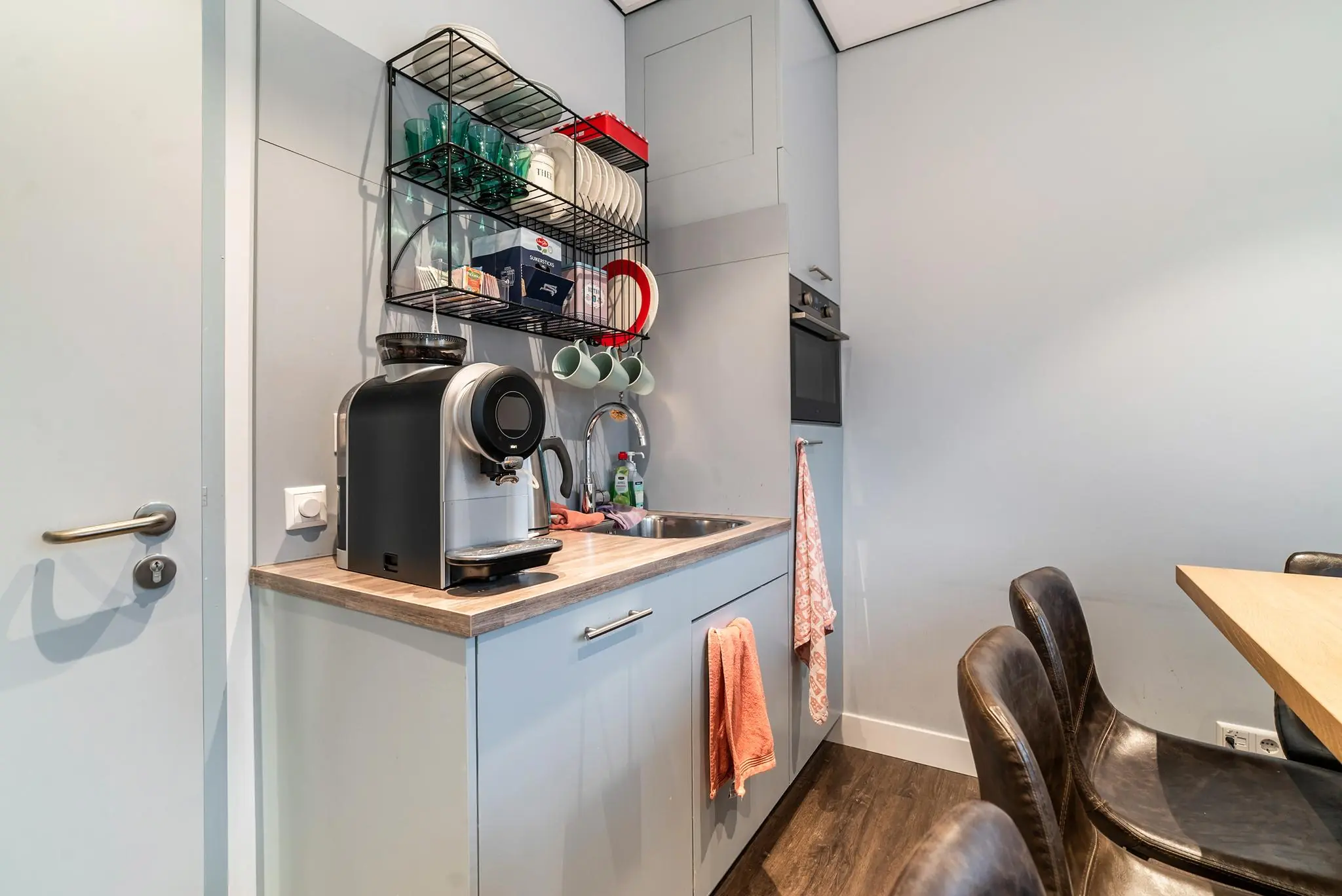 Moderne kitchenette aan de Bordewijklaan met koffiezetapparaat, spoelbak, oven en open wandrek met servies.