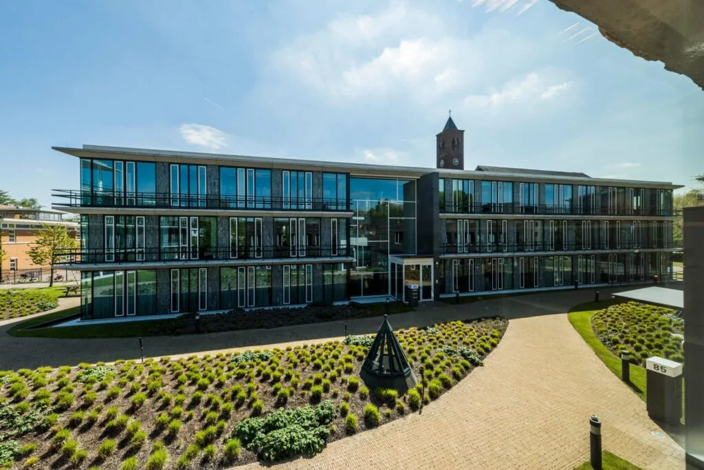Modern kantoorgebouw aan de De Cuserstraat 85 met glazen gevel en verzorgde tuin op de voorgrond.