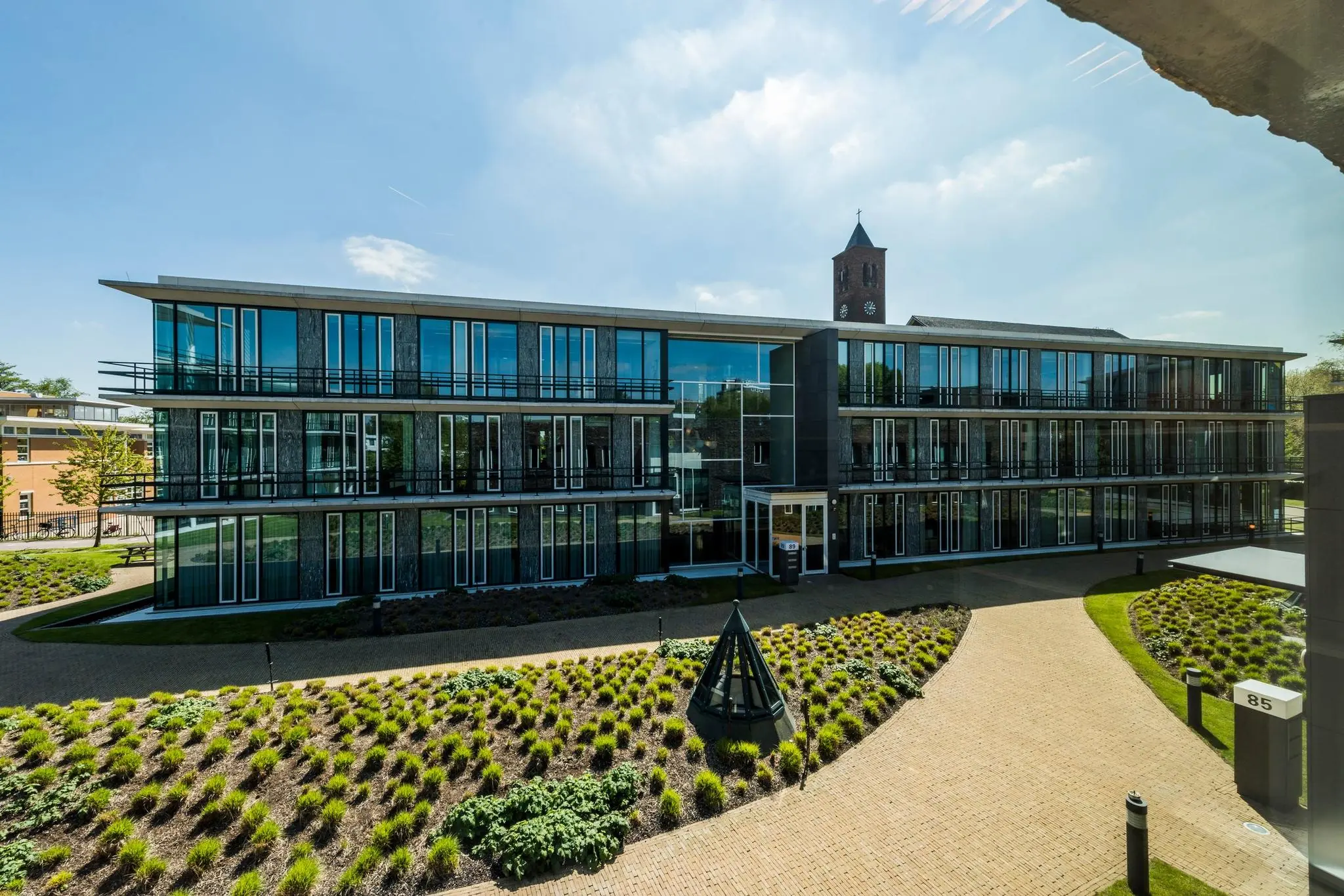 Modern kantoorgebouw aan de De Cuserstraat 85 met glazen gevel en verzorgde tuin op de voorgrond.