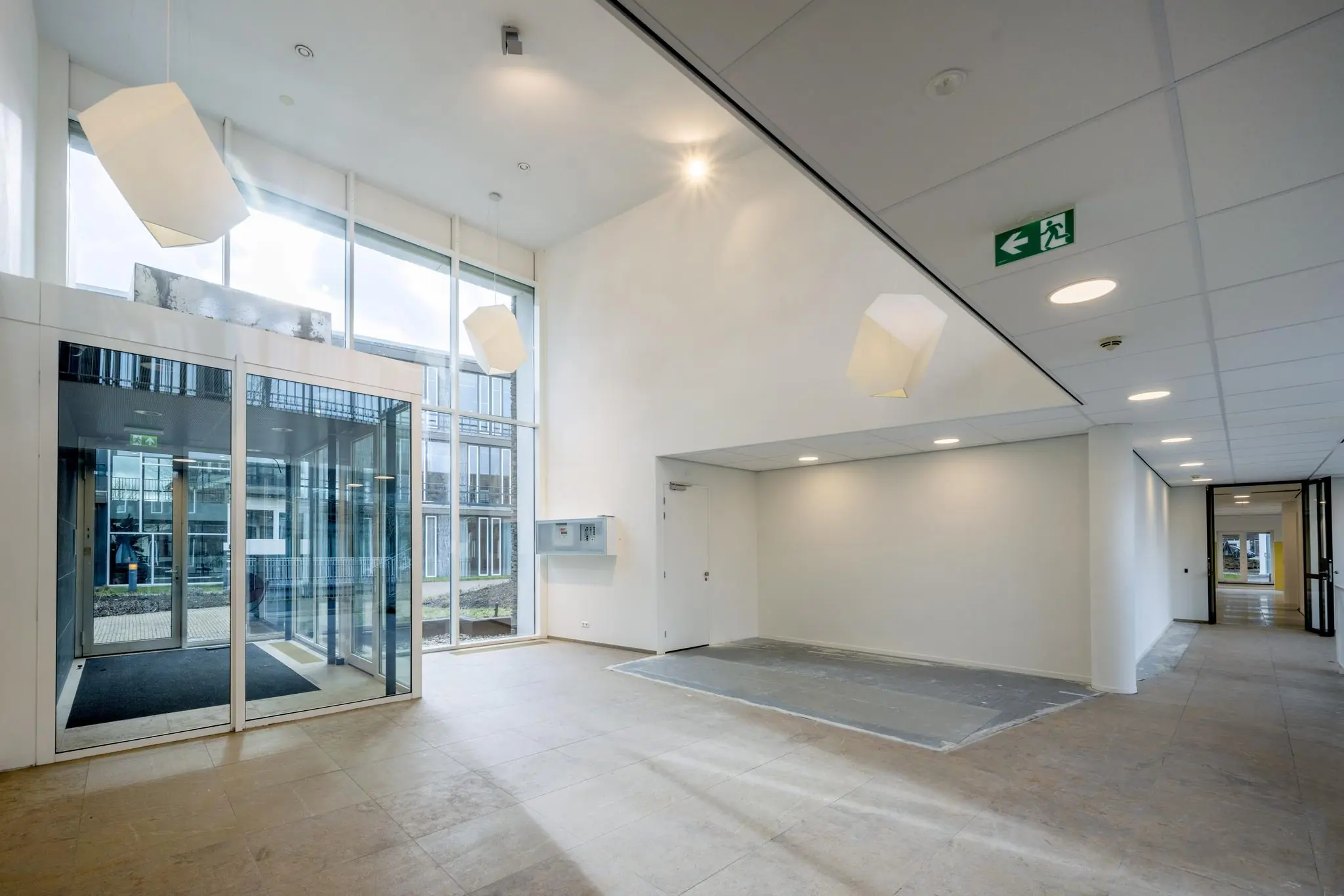 Moderne, lichte entreehal van een gebouw aan de De Cuserstraat met grote ramen en geometrische hanglampen.